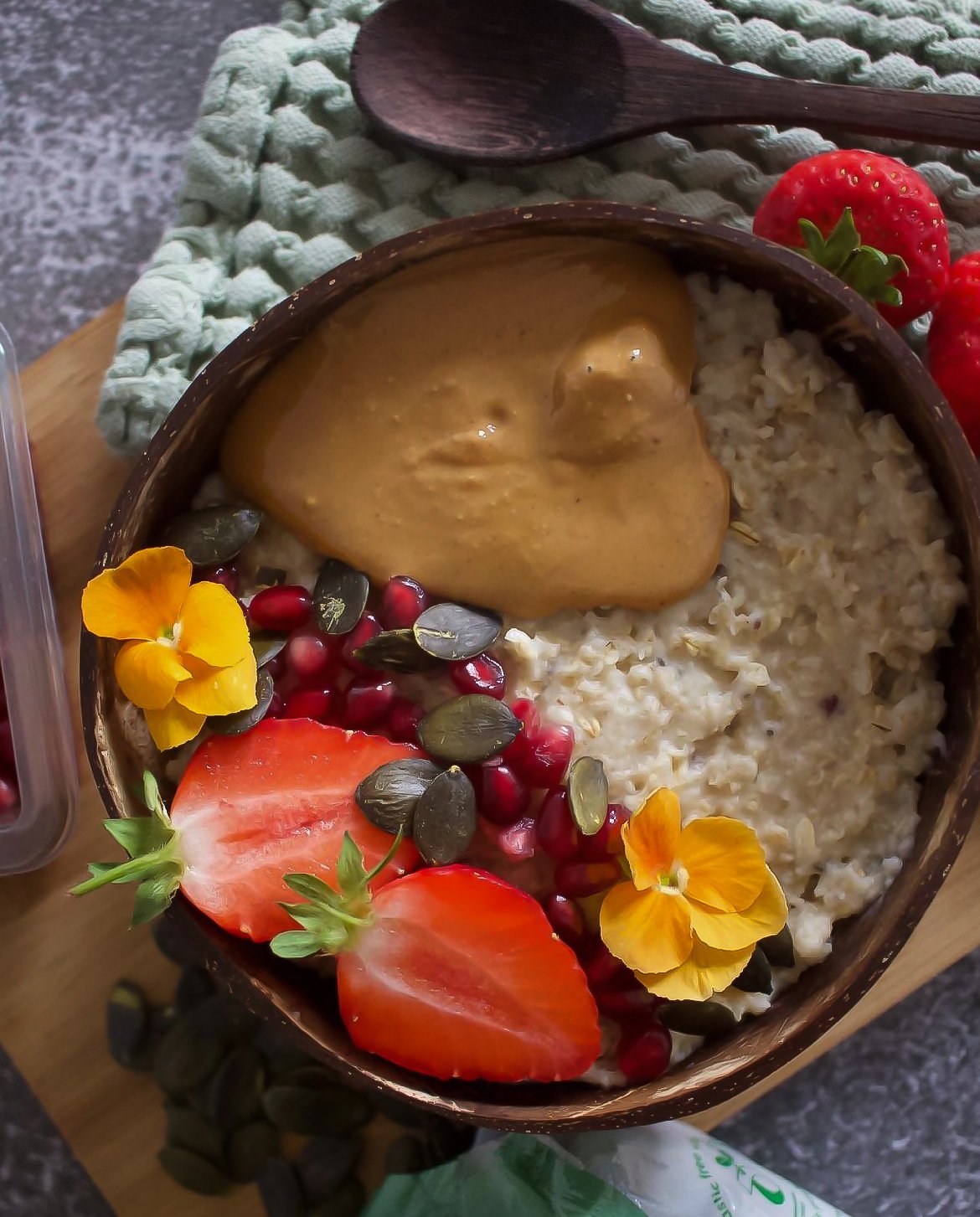 A bowl of oatmeal topped with peanut butter, sliced strawberries, pomegranate seeds, pumpkin seeds, and edible flowers, on a wooden surface with a spoon and additional strawberries nearby.