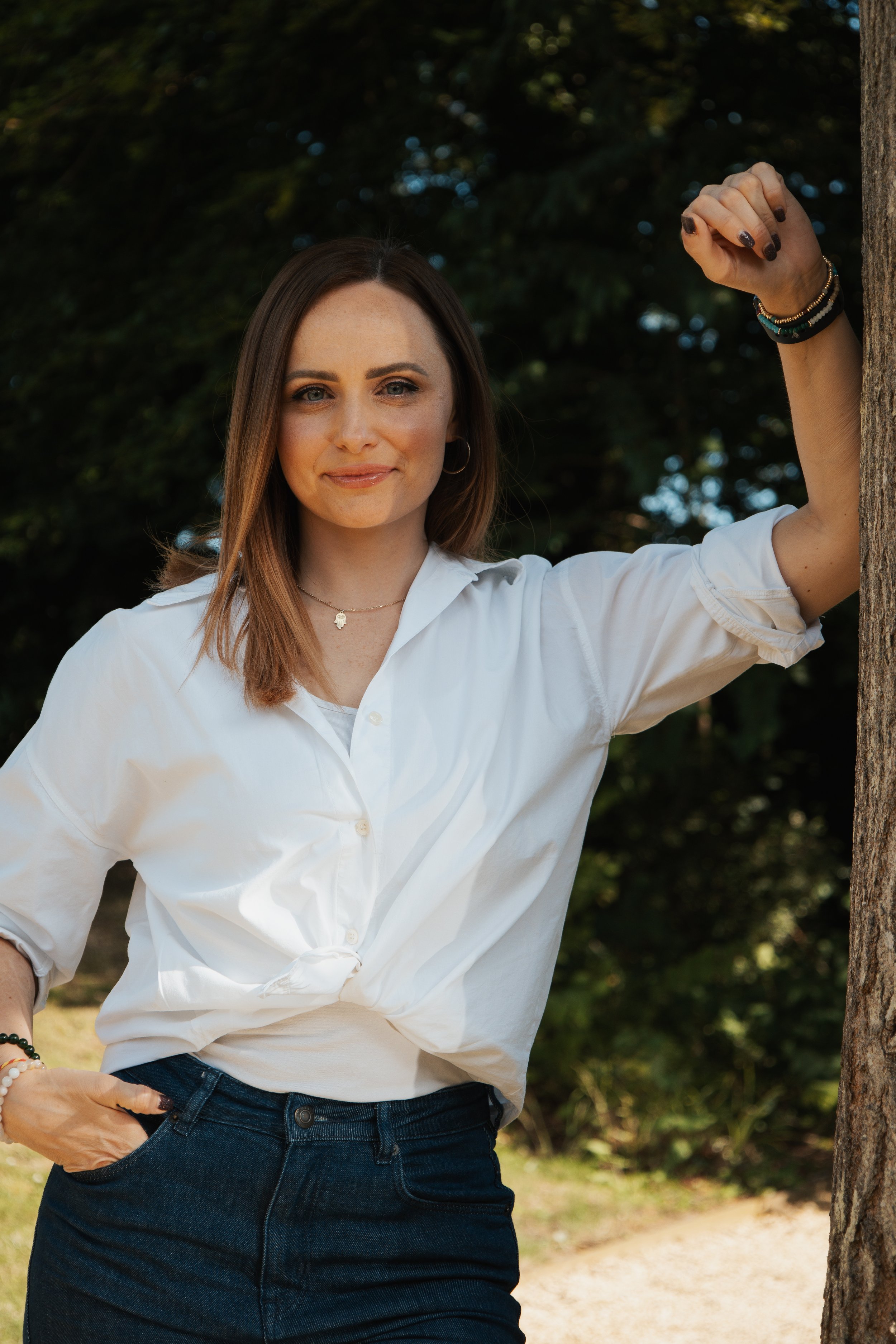 A woman with shoulder-length brown hair wearing a white shirt and dark jeans, standing outdoors near a tree, with a background of leafy trees and sunlight filtering through.