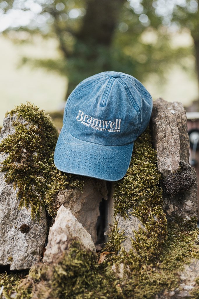 A blue baseball cap with 'Bramwell PRIME PROPERTY SEARCH' embroidered on it, placed on moss-covered rocks outdoors.