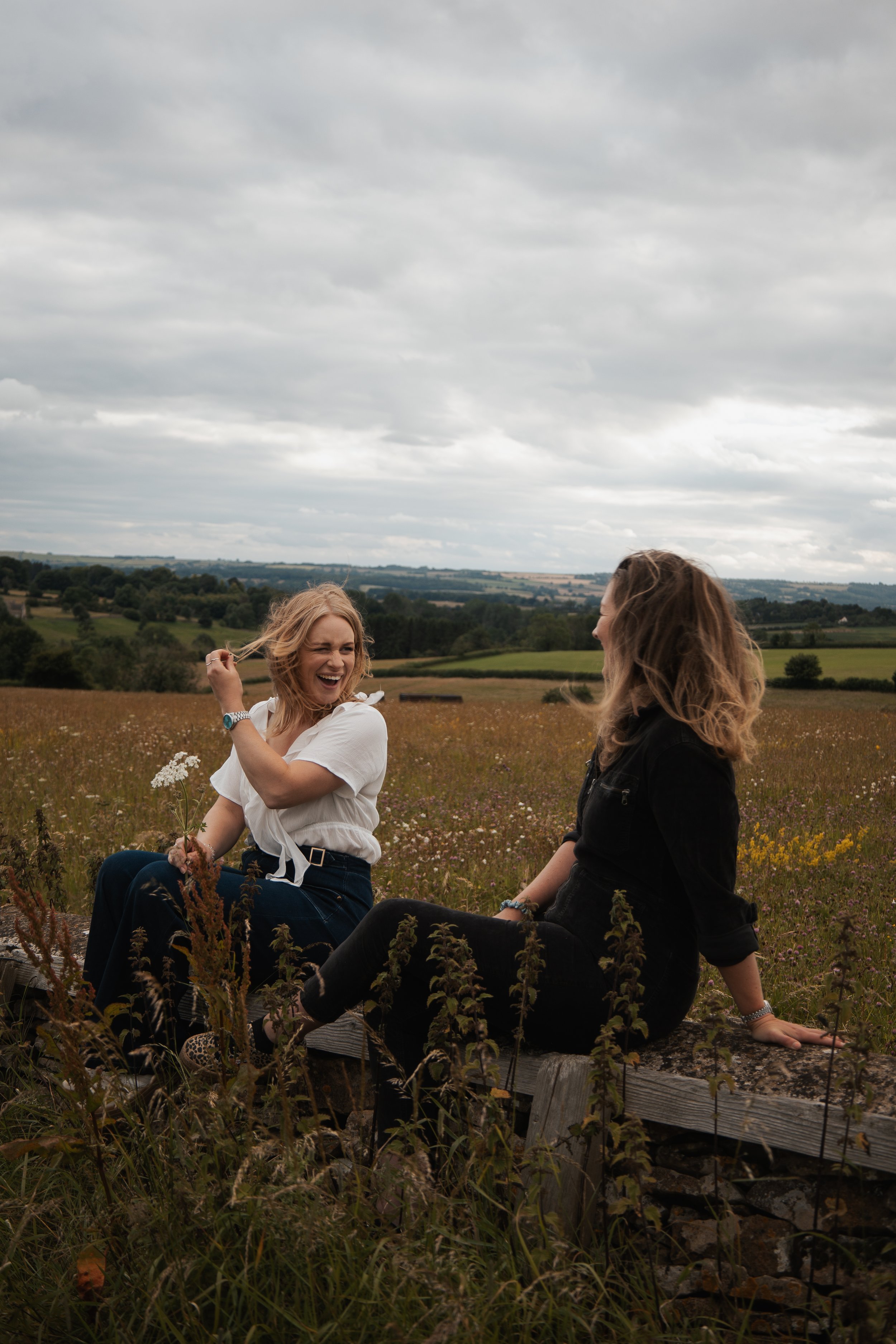Two women laughing and talking while sitting on a wooden bench in a field with wildflowers, overcast sky in the background.