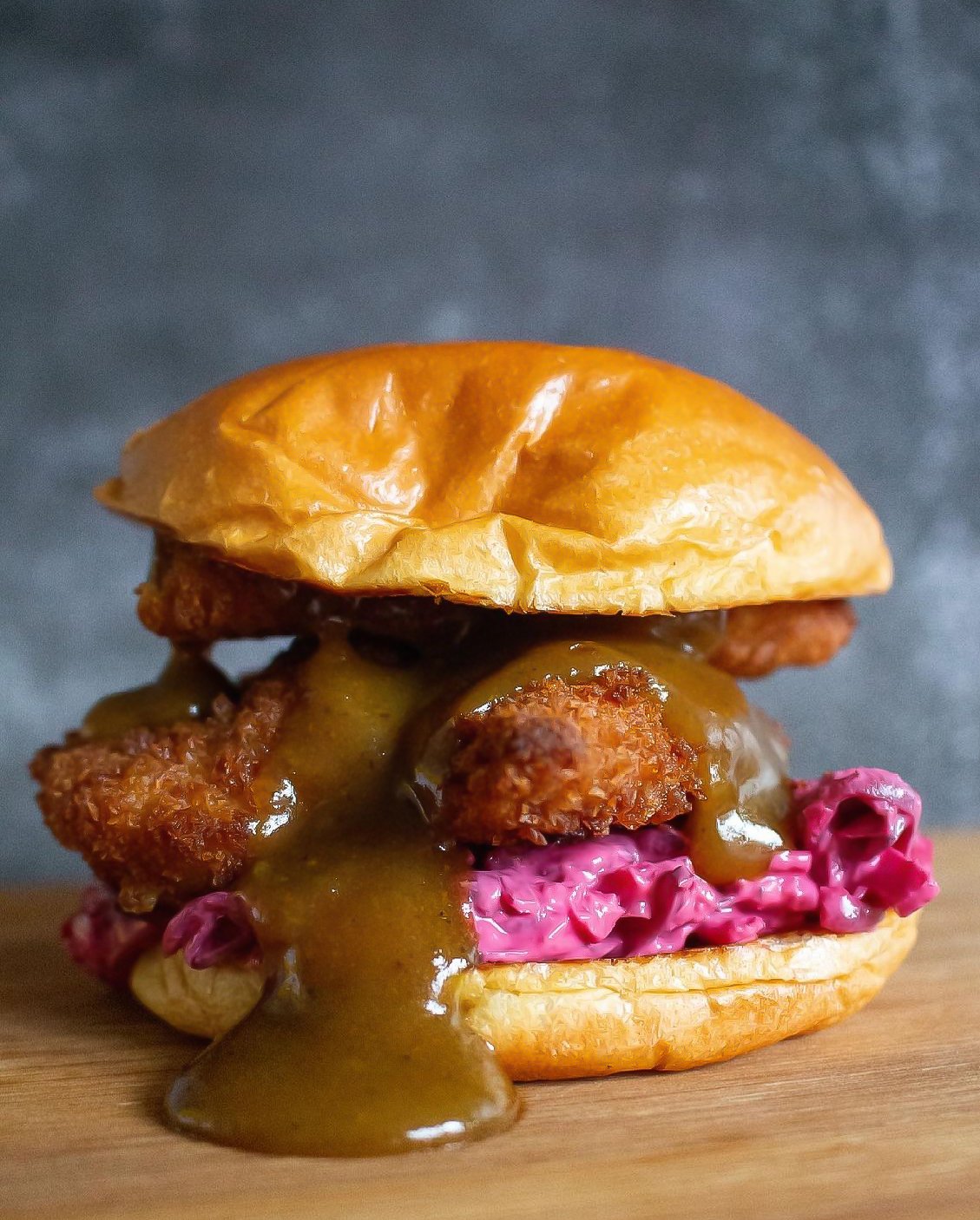 A close-up of a burger with a bun, purple coleslaw, fried chicken fillet, melted cheese, and a thick brown sauce topping.