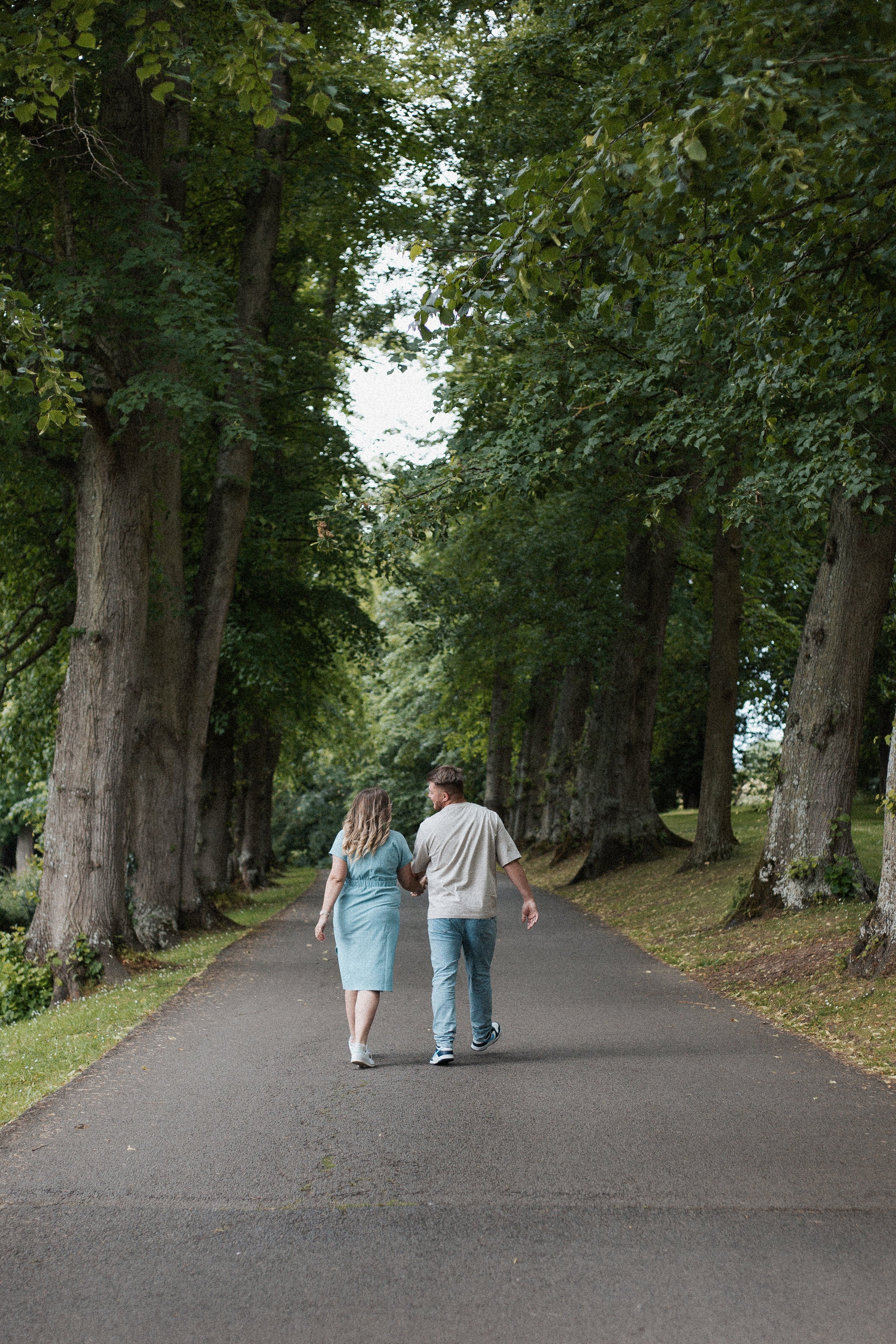 A couple walking along a tree-lined paved path in a park or forest, holding hands and engaging in conversation.