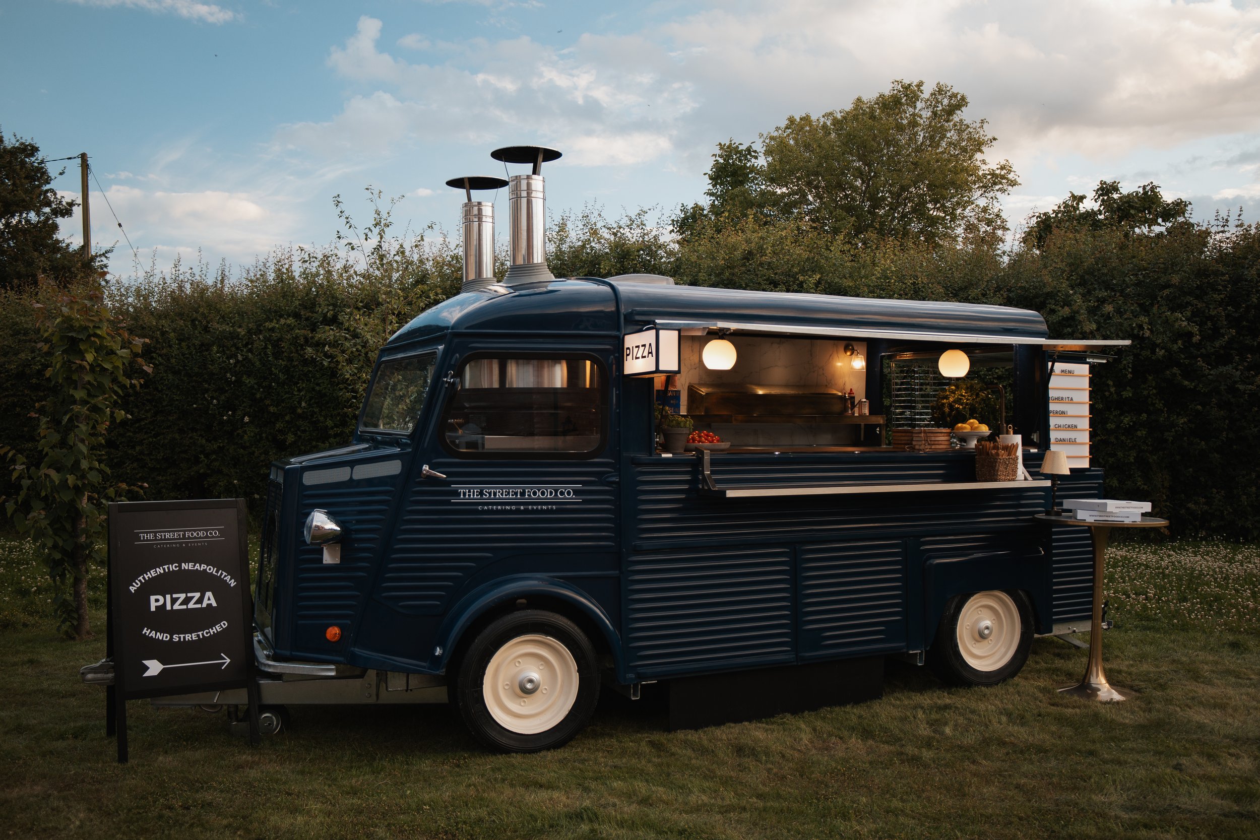 A vintage blue food truck serving pizza parked on grass with a sign indicating it as an authentic Neapolitan pizza stand.