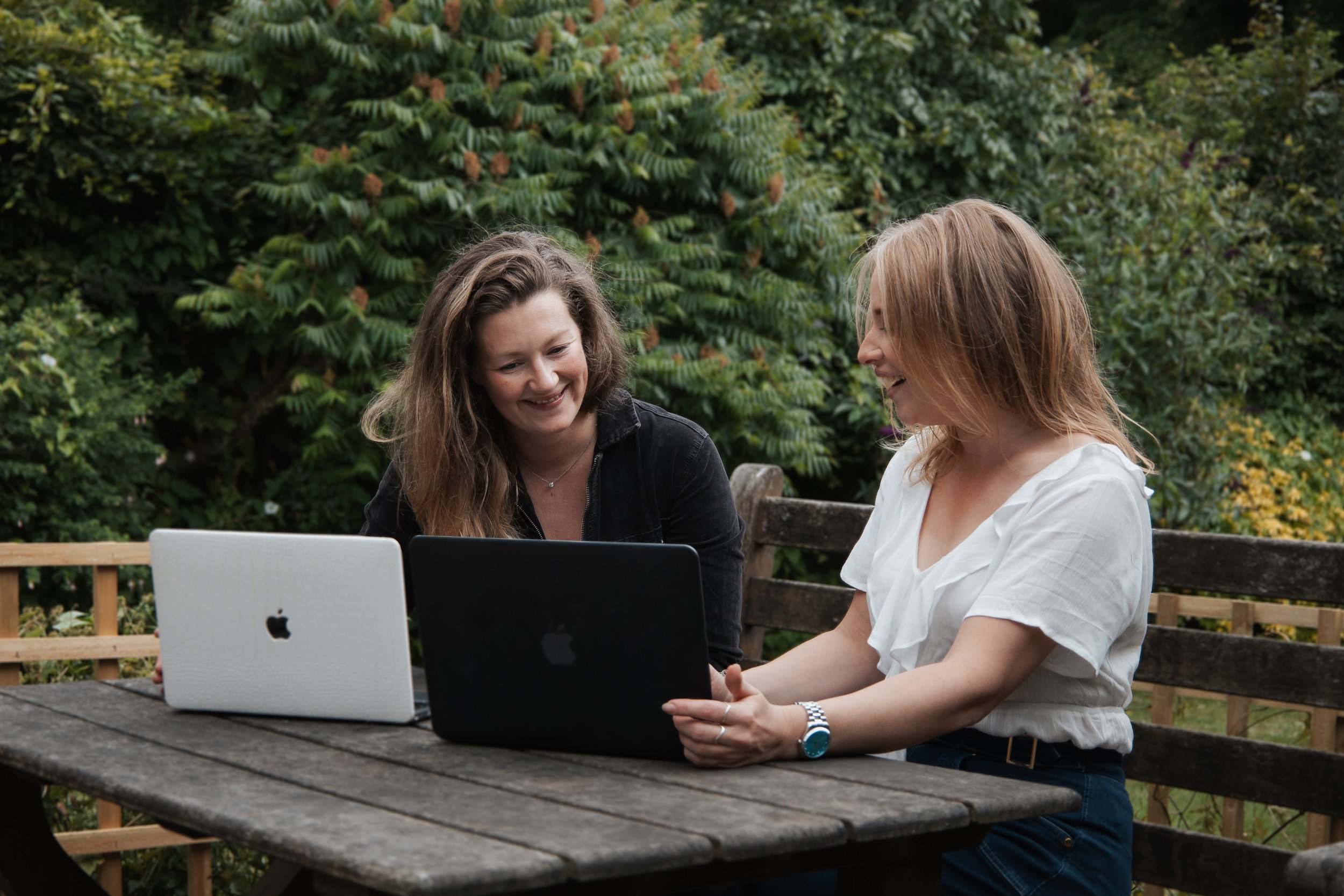 Two women sitting at a wooden outdoor table, smiling and looking at a black laptop, with another silver laptop on the table, surrounded by green trees.