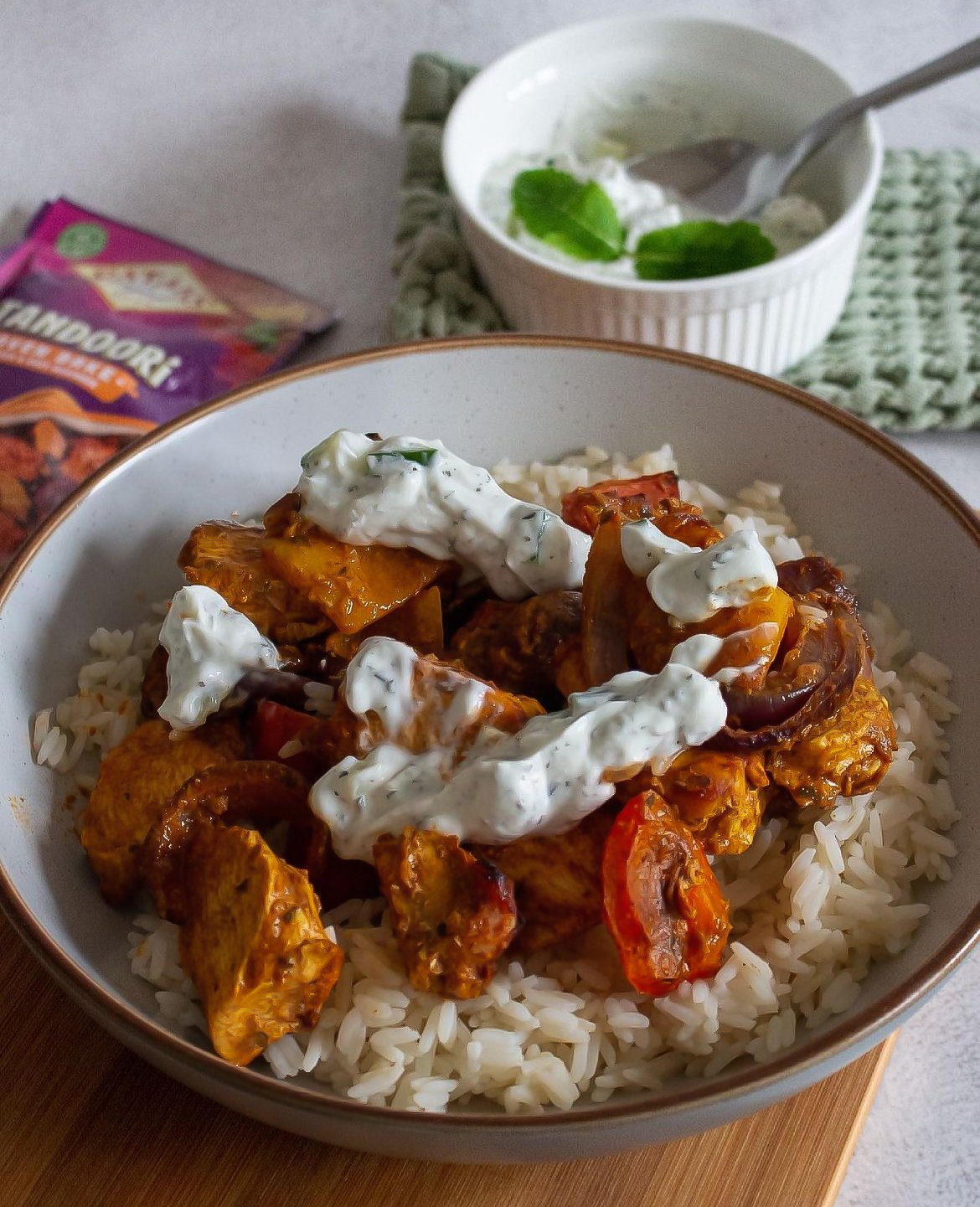 A bowl of cooked white rice topped with chicken, bell peppers, and a creamy white sauce, with a side of cucumber and herbs in a white ramekin and a packet of seasoning in the background.