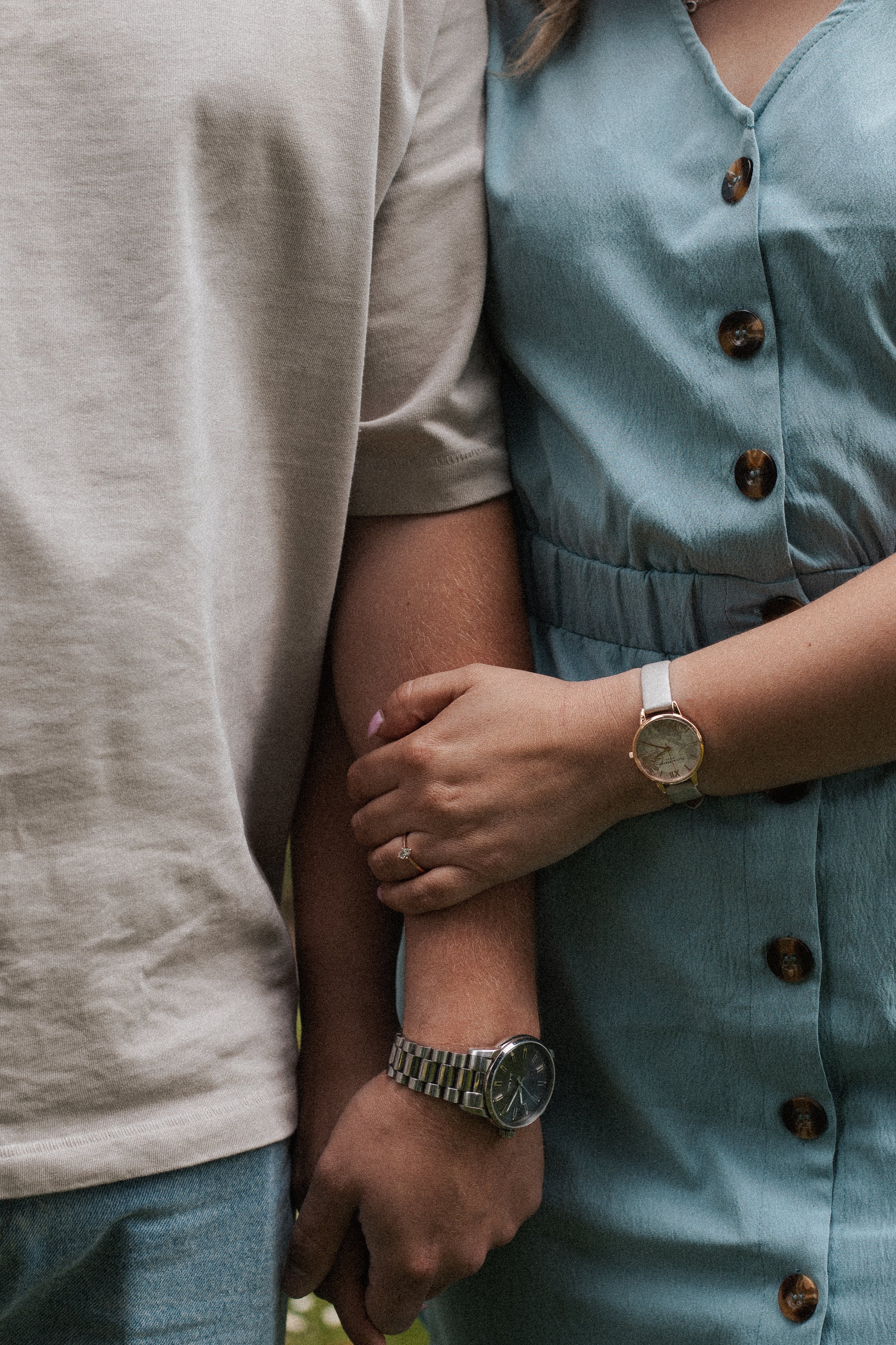 A close-up of two people holding hands, with one person's arm around the other. The person on the right is wearing a blue dress with buttons and a watch, while the other person is wearing a beige shirt and a watch.