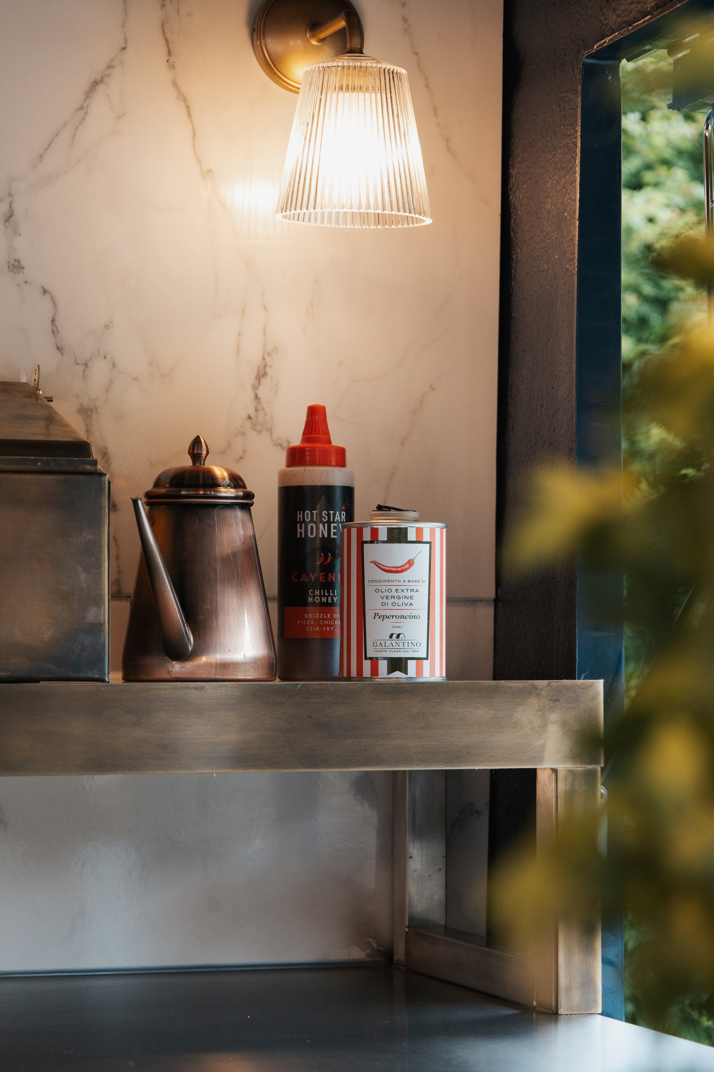 A kitchen shelf with a metal teapot, a bottle of hot honey, and a can of olive oil, illuminated by a wall-mounted light fixture, with a window showing greenery outside.