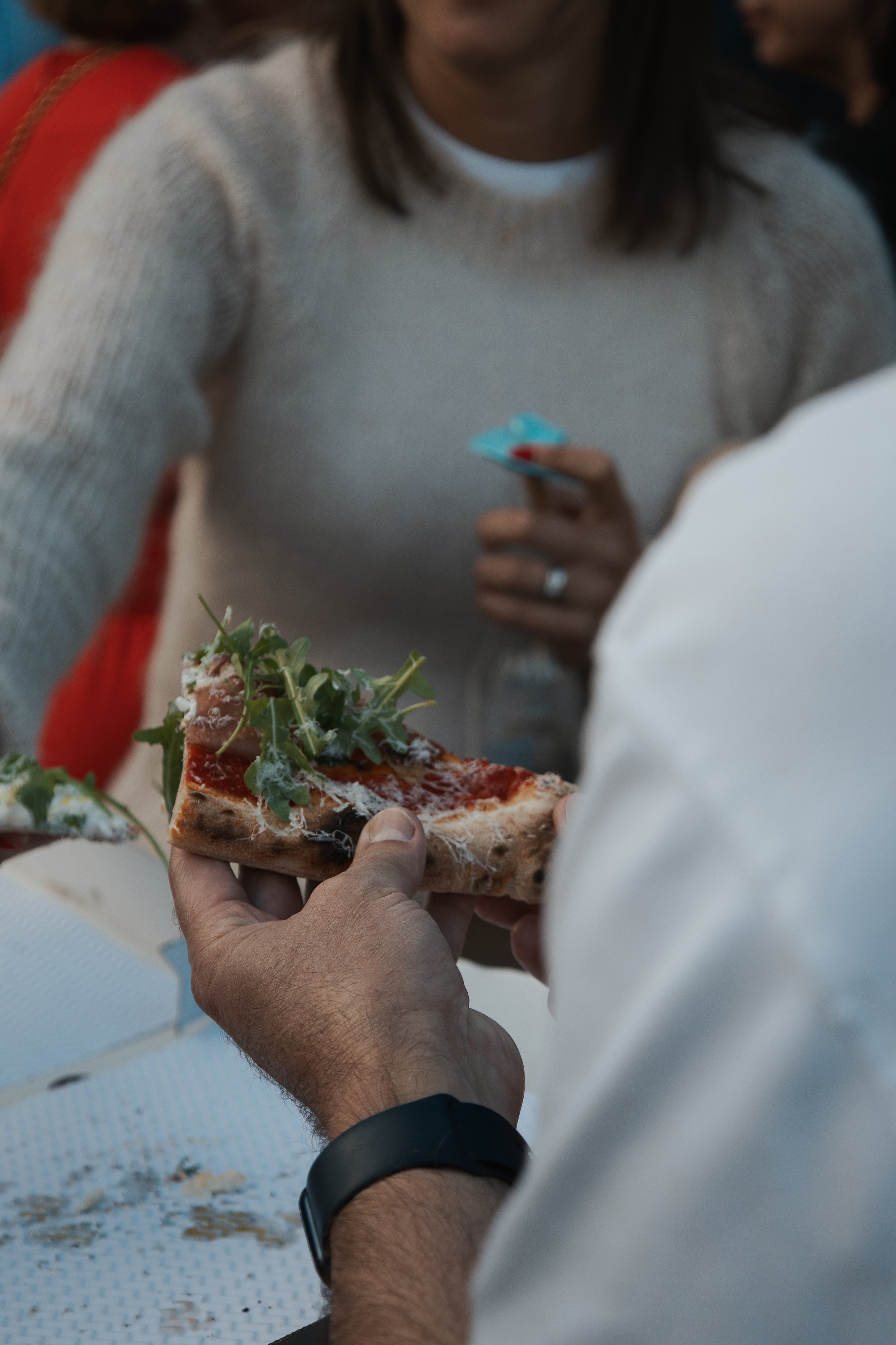 Close-up of a person holding a slice of pizza with arugula and cheese, with another person in the background wearing a white sweater, holding a credit card and a water bottle.