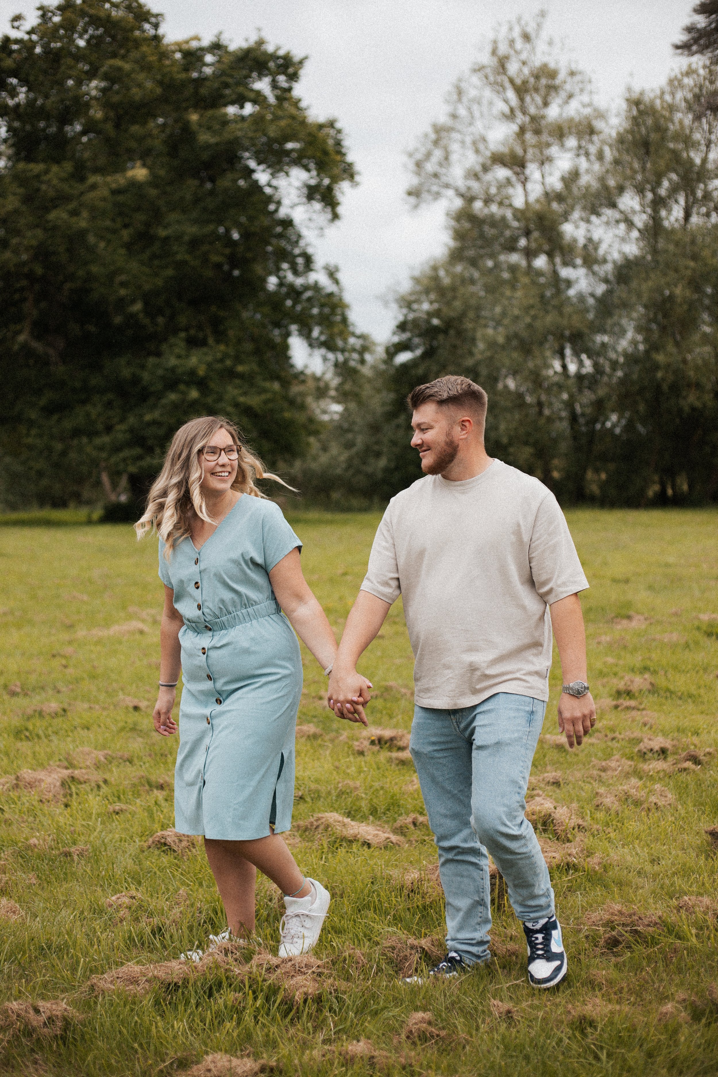 A smiling couple holding hands and walking through a grassy field, with trees in the background on an overcast day.