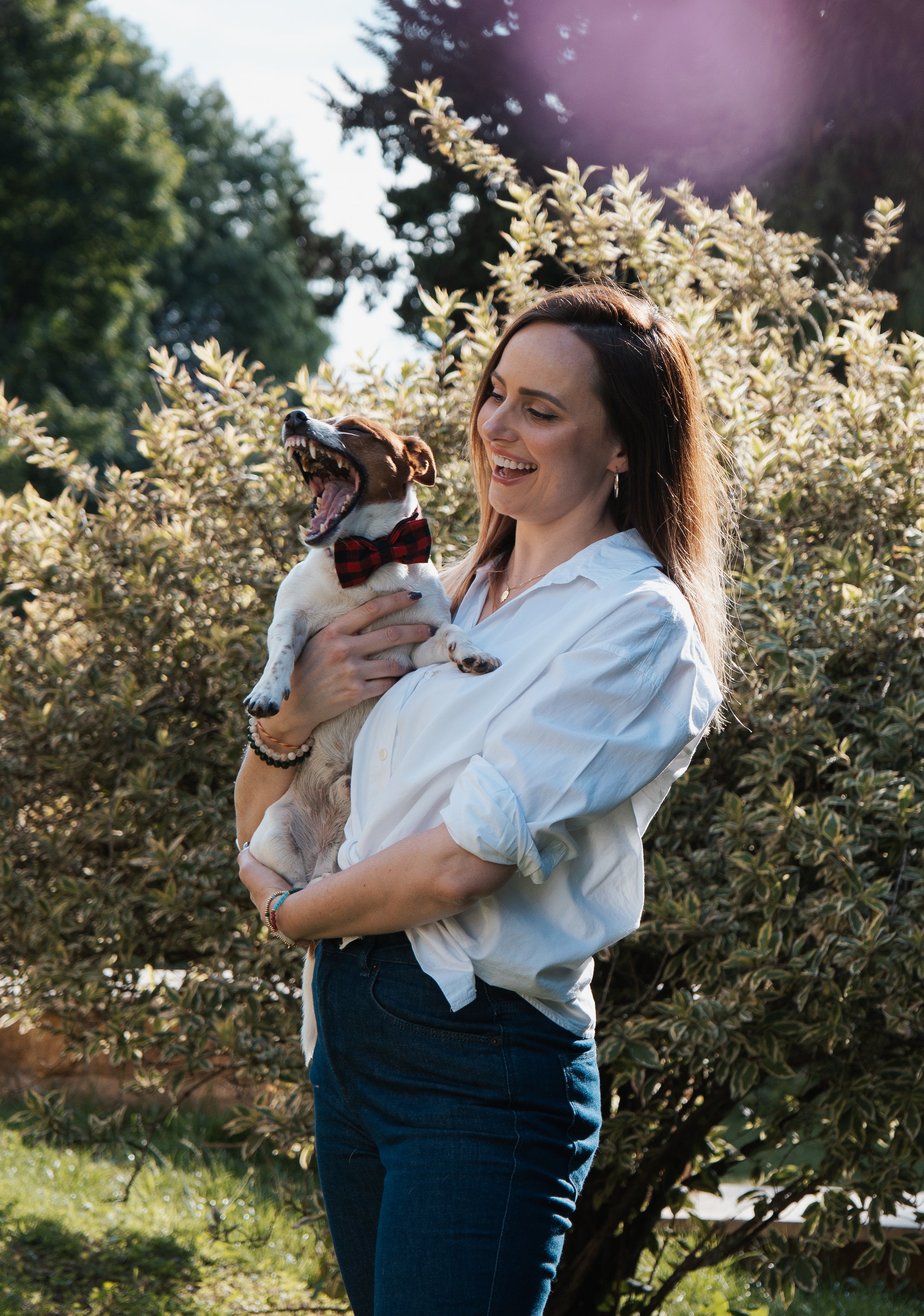 A woman in a white shirt holding a small dog with a red and black bow tie, standing outdoors with trees and bushes in the background, as the dog yawns.