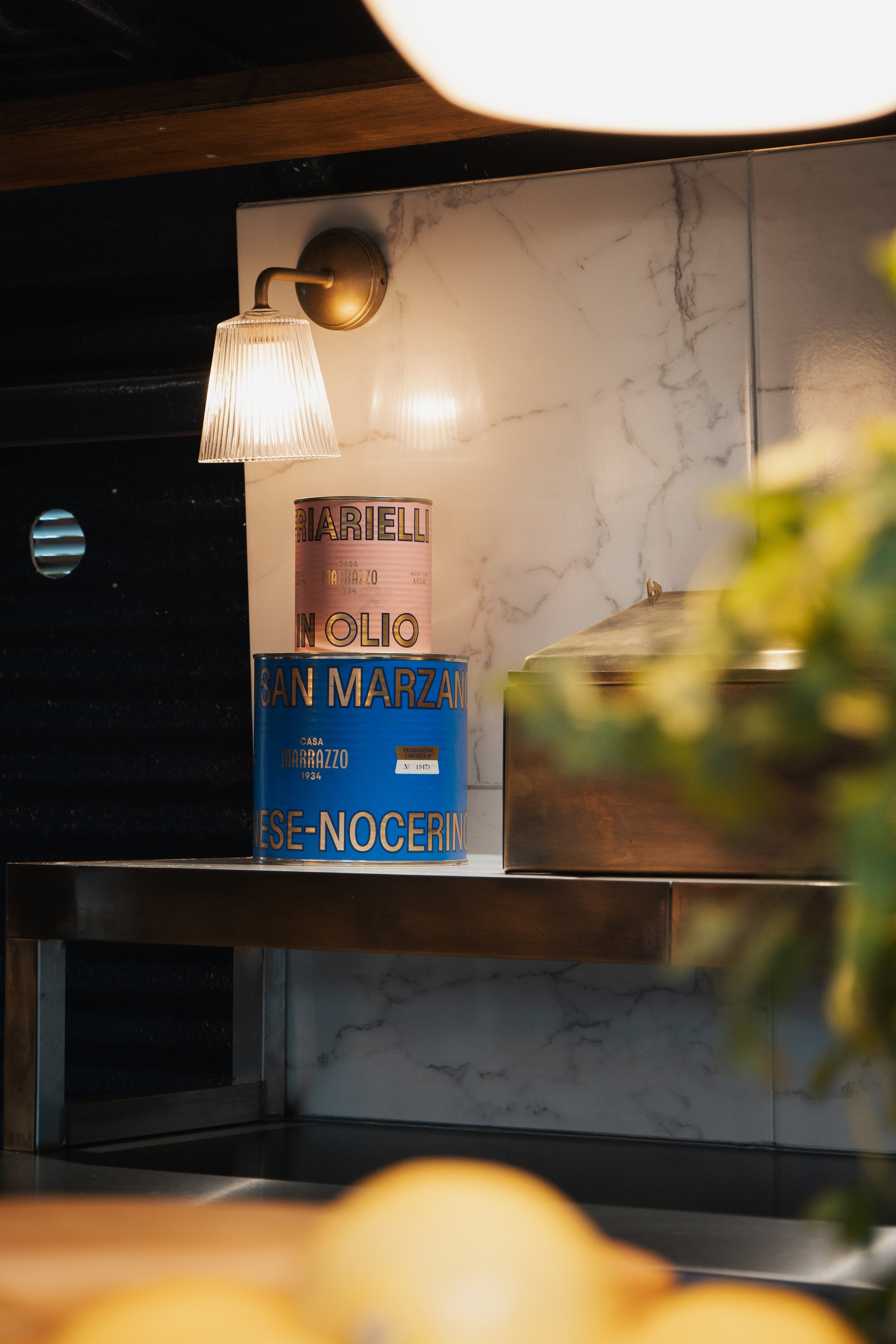 Decorative cans of Marzotto pasta on a kitchen shelf illuminated by a wall-mounted lamp, marble backsplash, and a stainless steel box.