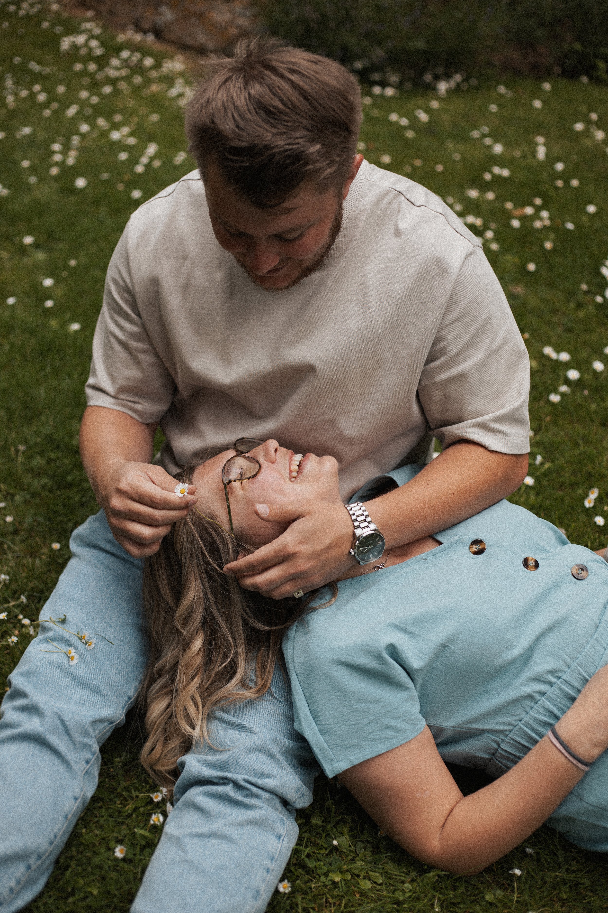 A man and woman lying on a grassy field, smiling and laughing while holding each other. The woman is on her back, and the man is leaning over her, playfully holding her glasses and smiling.