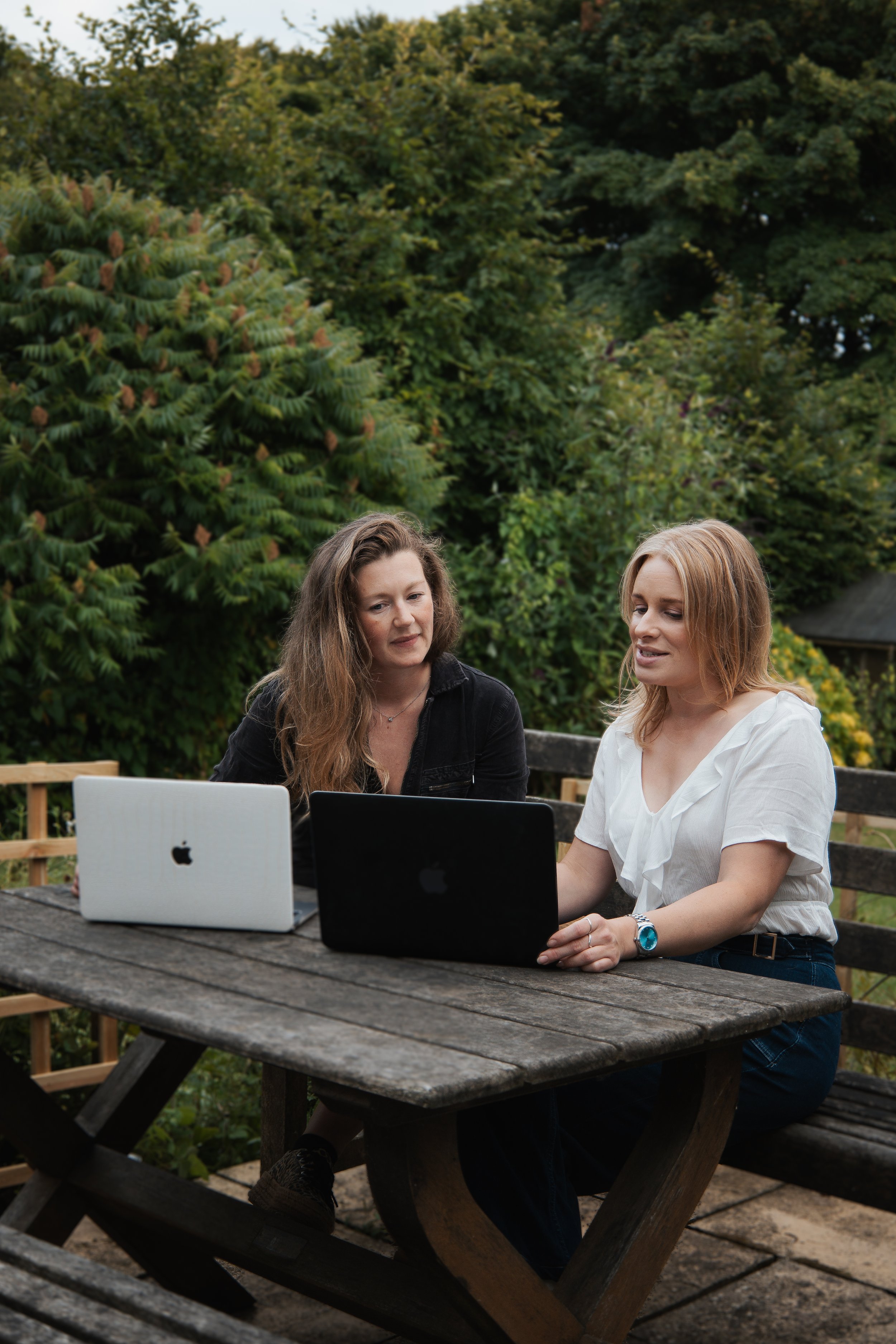 Two women sitting at a wooden outdoor table with laptops, discussing outdoors with green trees in the background.