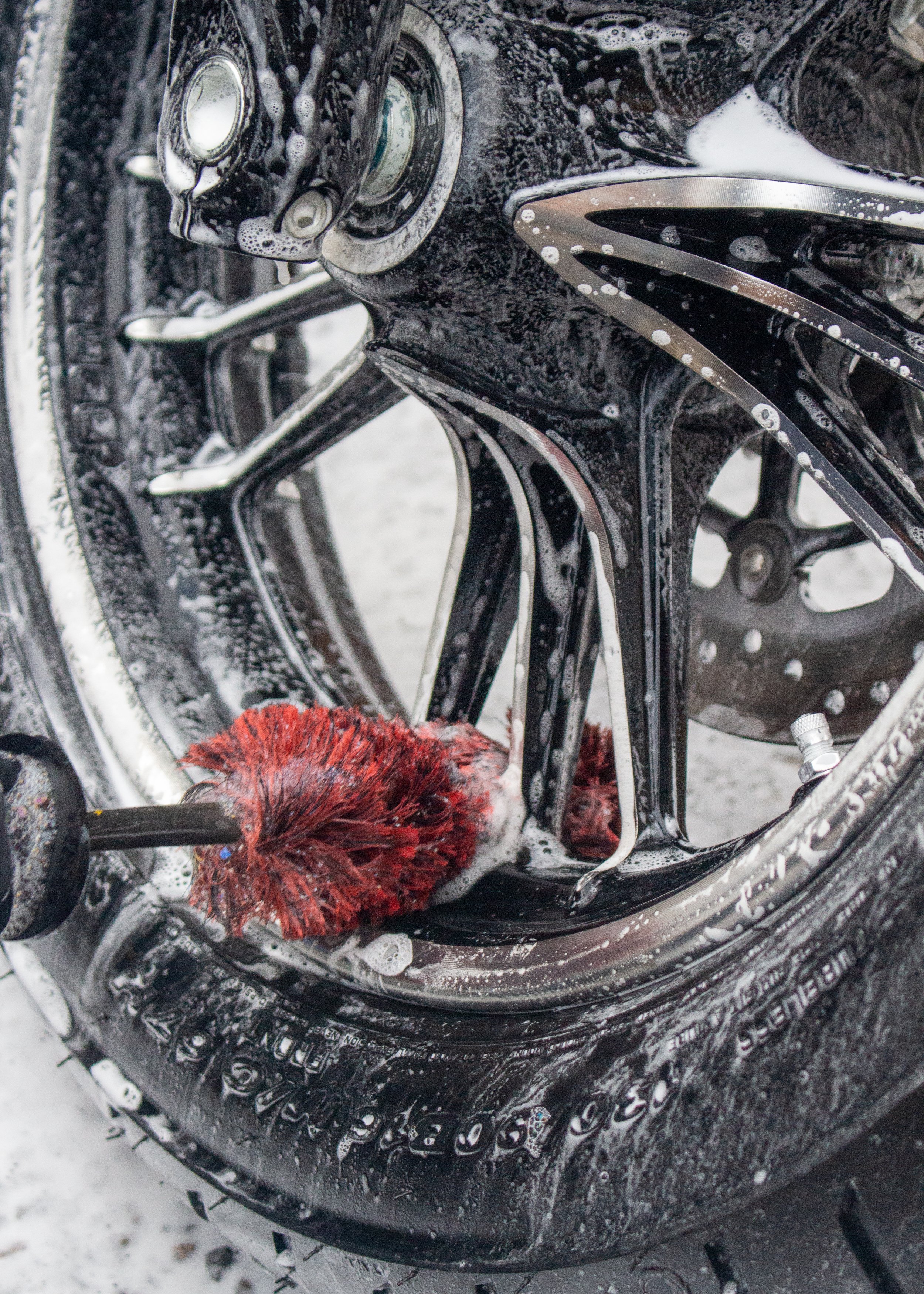 Close-up of a black bicycle frame being washed with soap and a red brush, covered in soap suds.