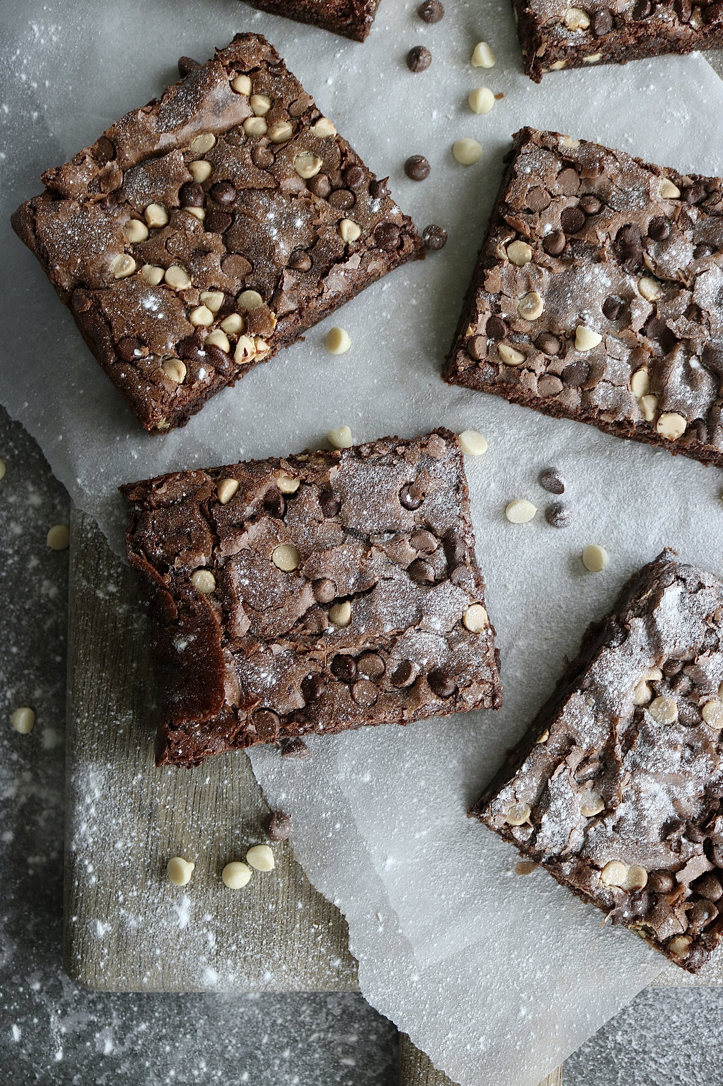 Four pieces of chocolate brownies topped with white and dark chocolate chips, on a parchment-lined wooden board sprinkled with powdered sugar.