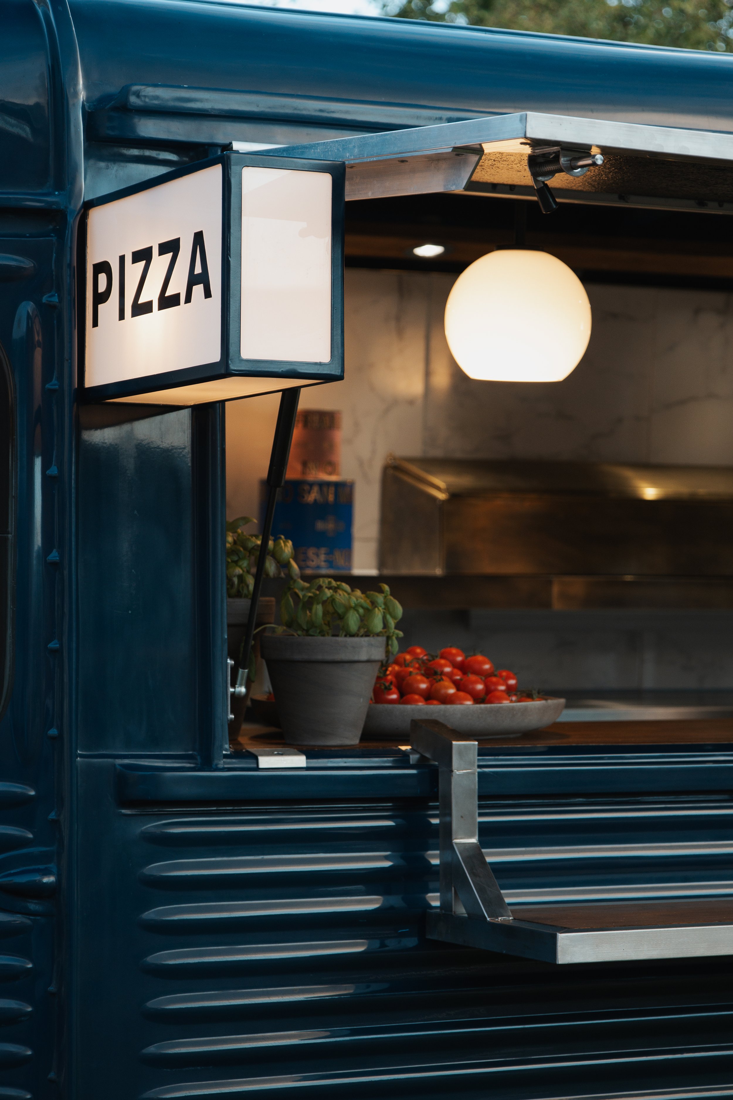 A food stand with a white sign labeled 'PIZZA', a bowl of cherry tomatoes, a potted basil plant, and a hanging round light bulb inside.