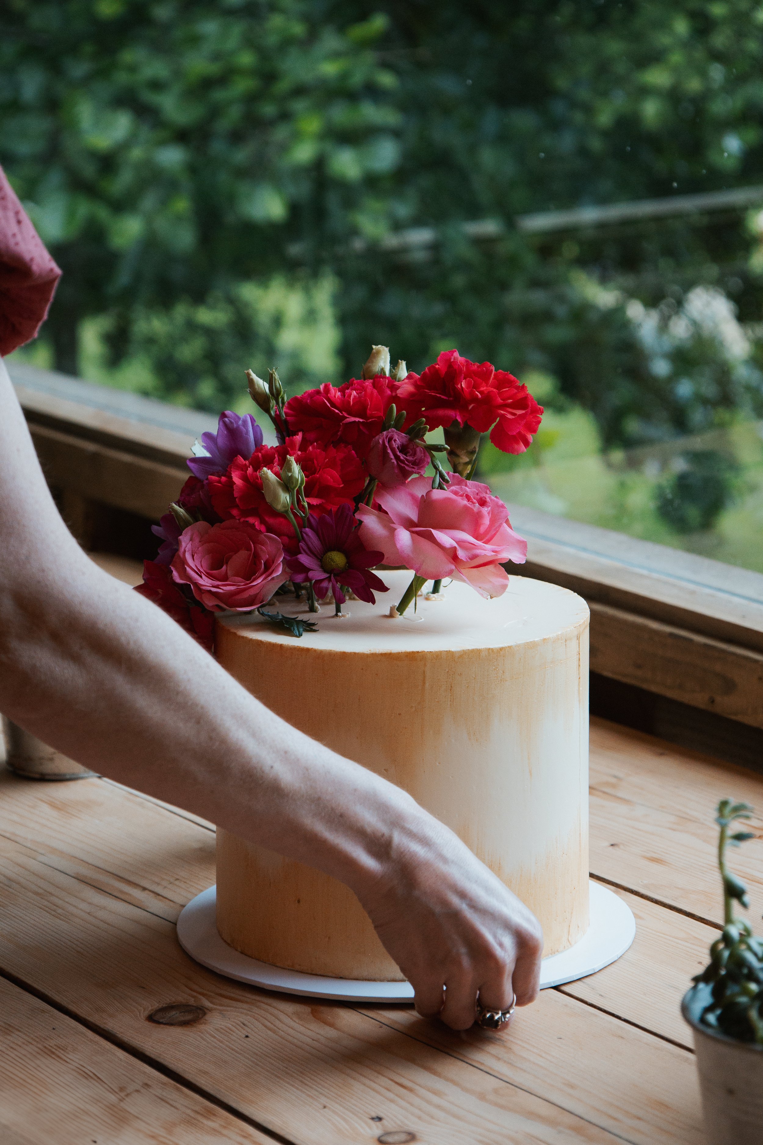 A person arranging a floral cake decoration on a wooden table with a window and green trees in the background.