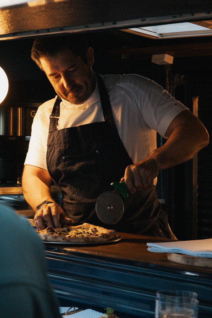 A chef in a black apron and white shirt slicing a pizza with a pizza cutter in a dimly lit restaurant kitchen.