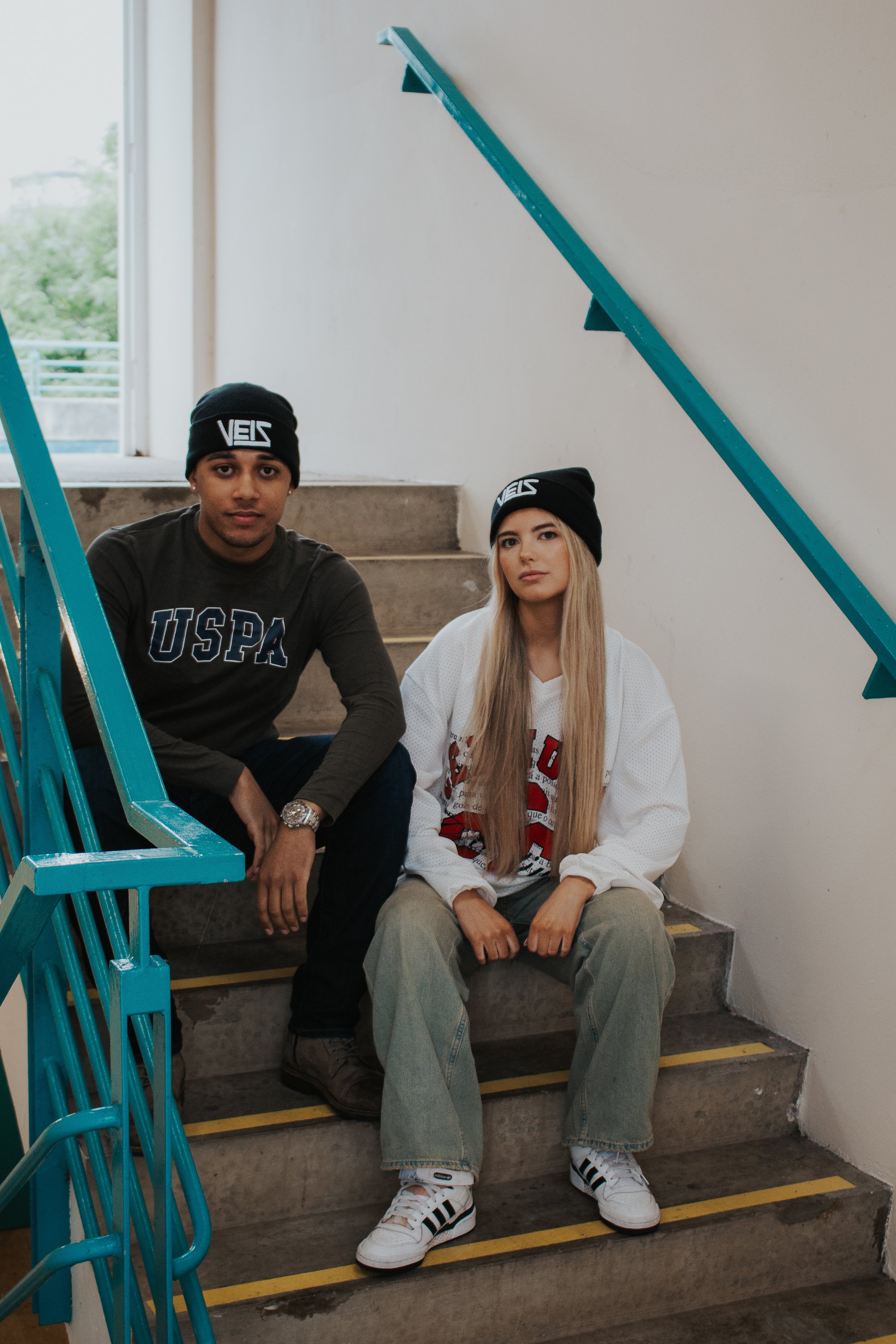 Two young adults sitting on stairs inside a building, both wearing black beanies with white lettering. The man on the left has a dark shirt with "USPA" printed on it, and the woman on the right has long blonde hair, a white sports jersey, and beige p
