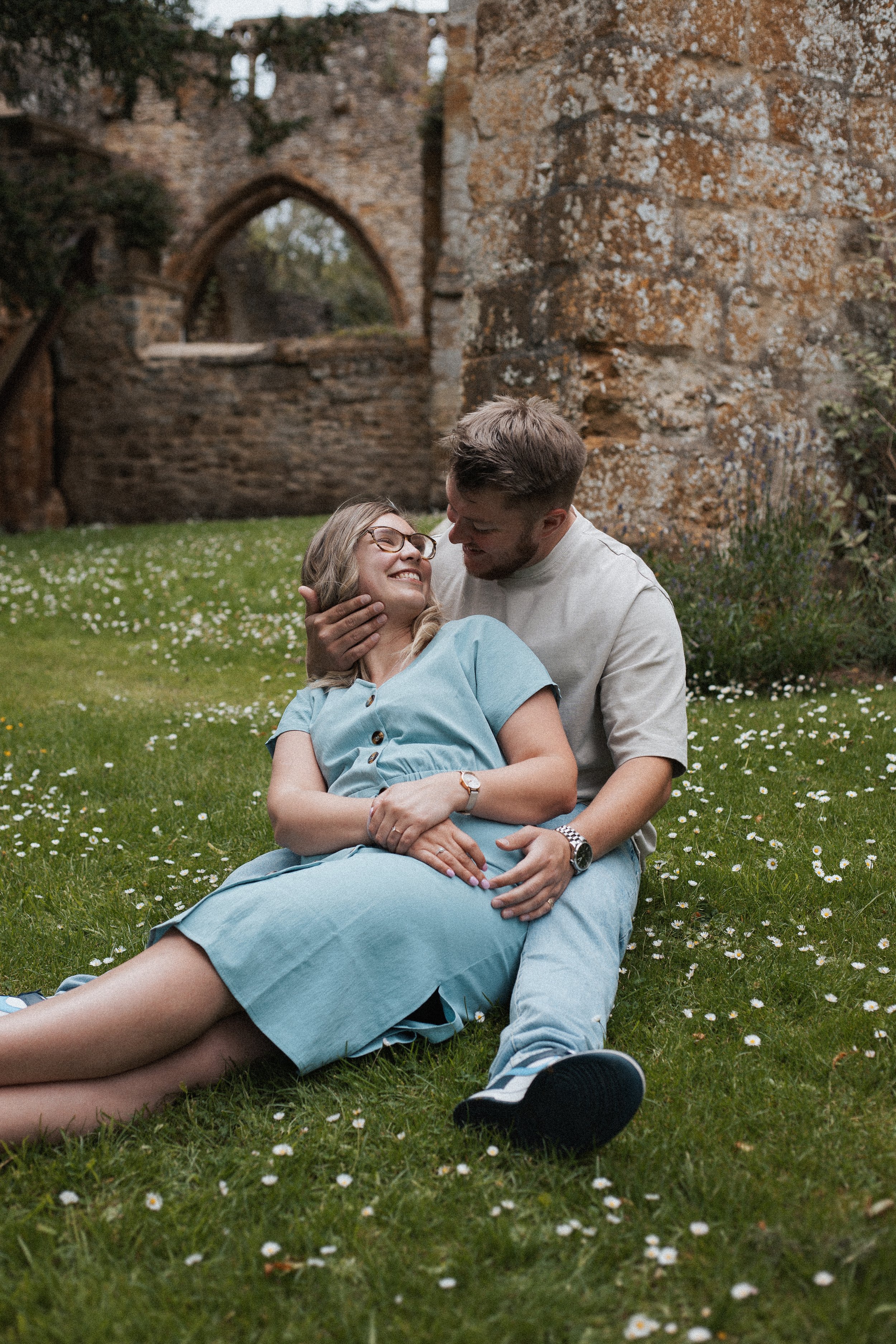 A couple sitting on the grass in front of a stone ruin, smiling and looking at each other affectionately. The woman sits with her legs extended and the man is holding her face, both enjoying a tender moment.