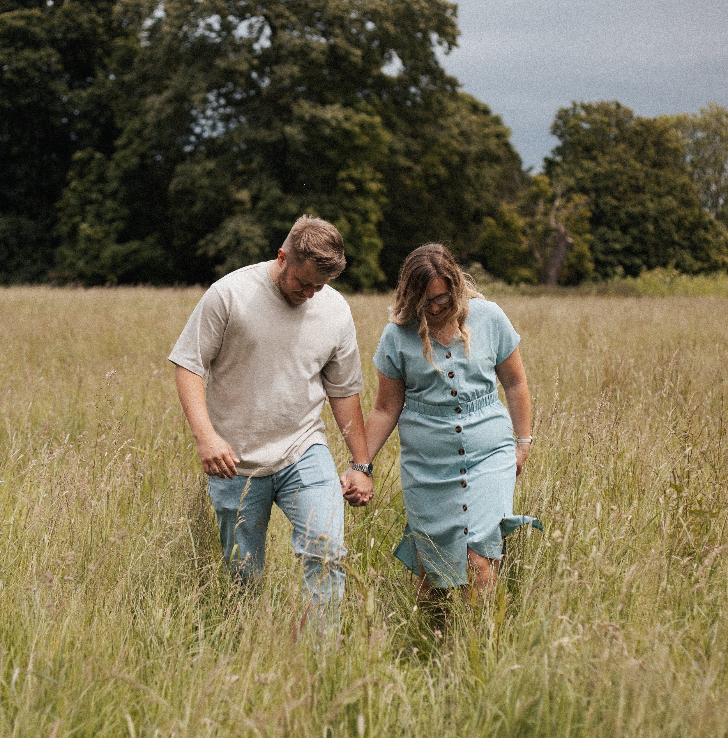 A couple walking hand-in-hand through a grassy field with trees in the background.