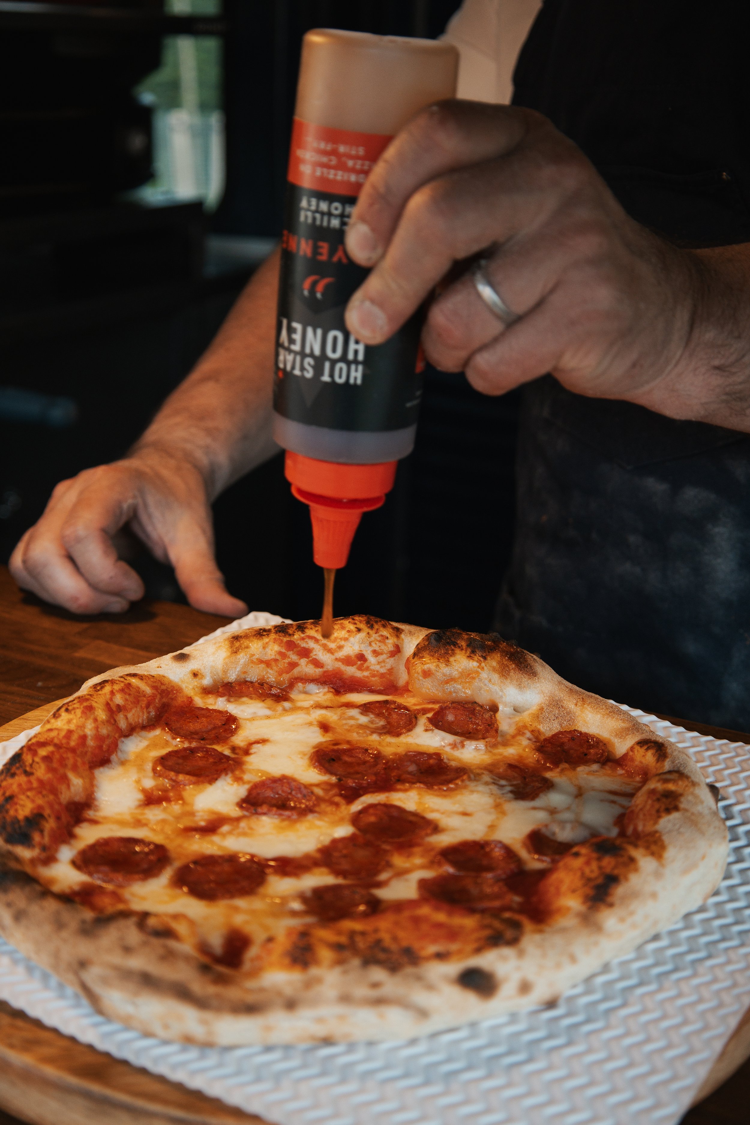Person squeezing hot sauce onto a pepperoni pizza with a golden-brown crust.
