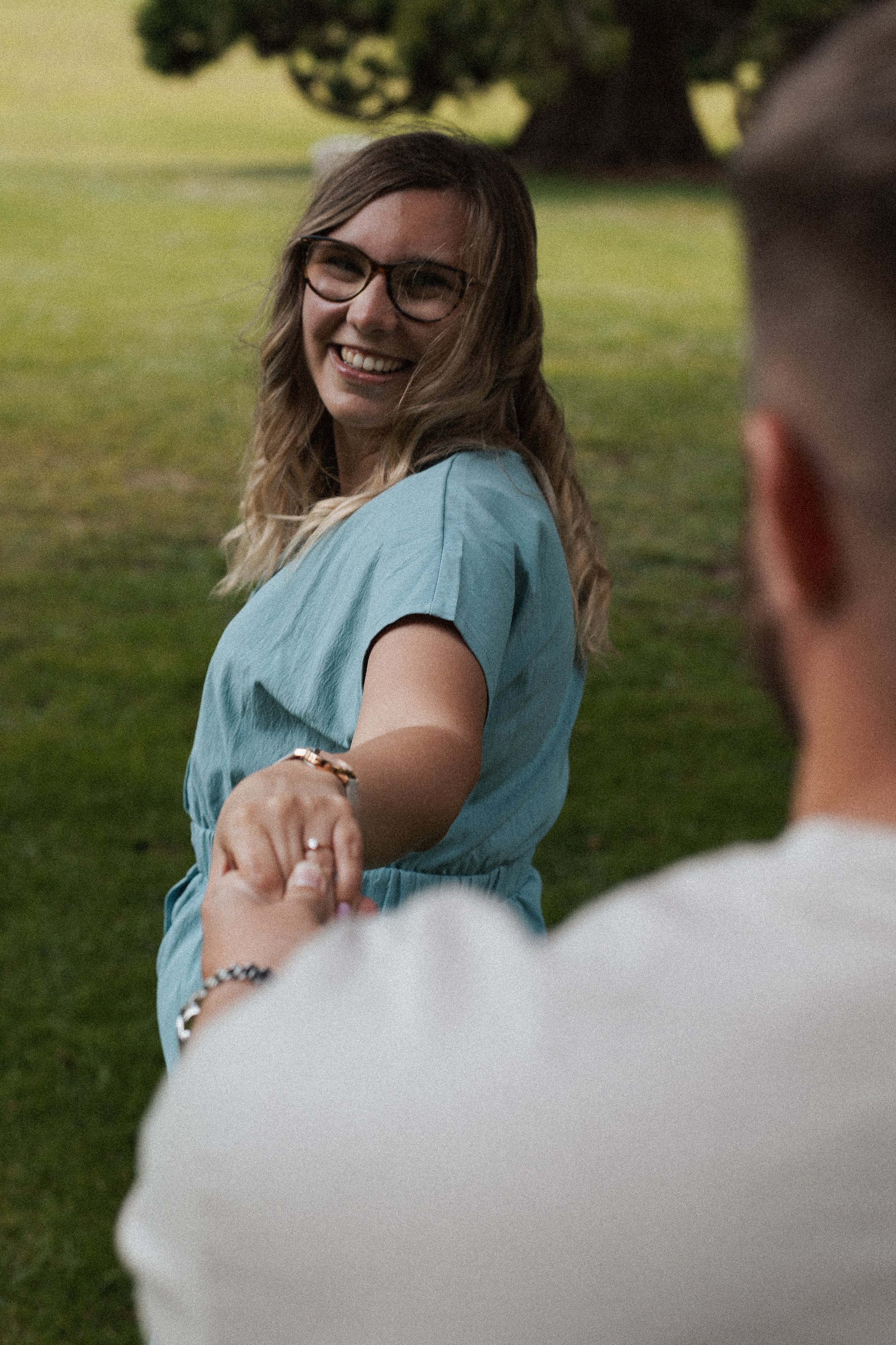 A woman with glasses, wavy hair, and a blue dress is smiling and holding hands with a man on a grassy field with trees in the background.