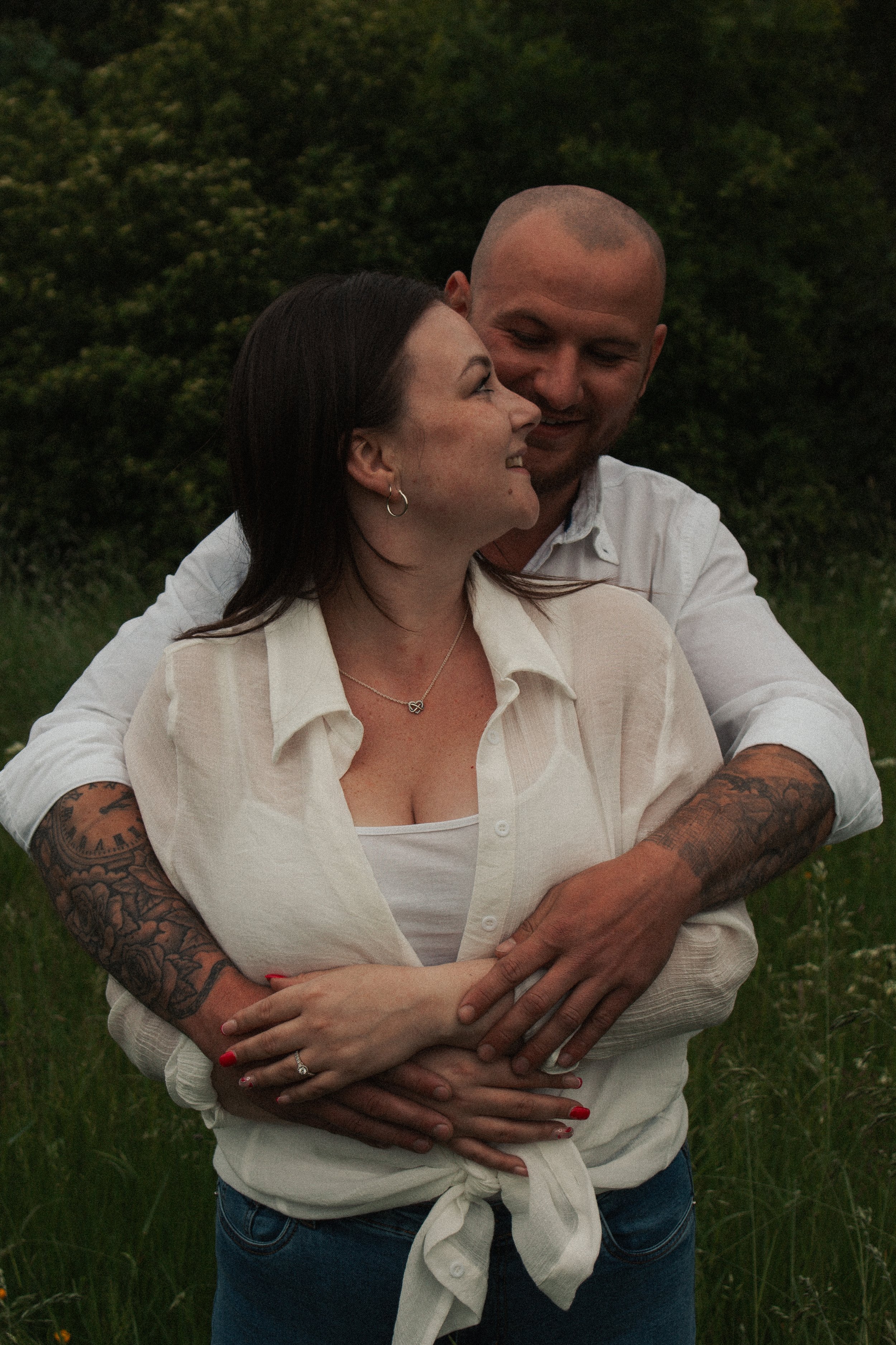 A couple is embracing outdoors in a grassy area with trees in the background, smiling and looking at each other affectionately.