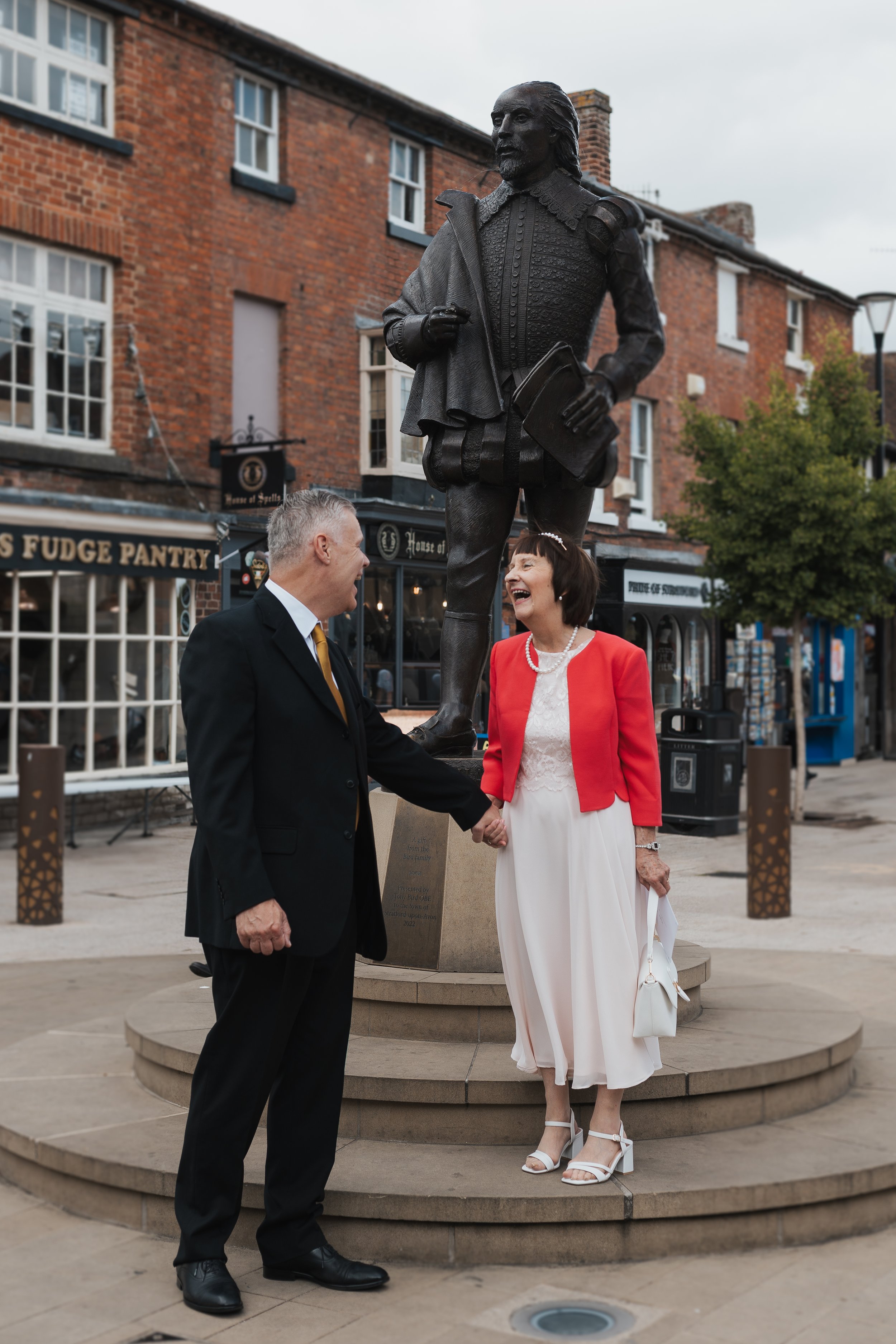A man and woman are holding hands and laughing in front of a statue of William Shakespeare in a town square. The man is wearing a black suit and tie, and the woman is wearing a white dress with a red jacket and white heels. The statue depicts a beard