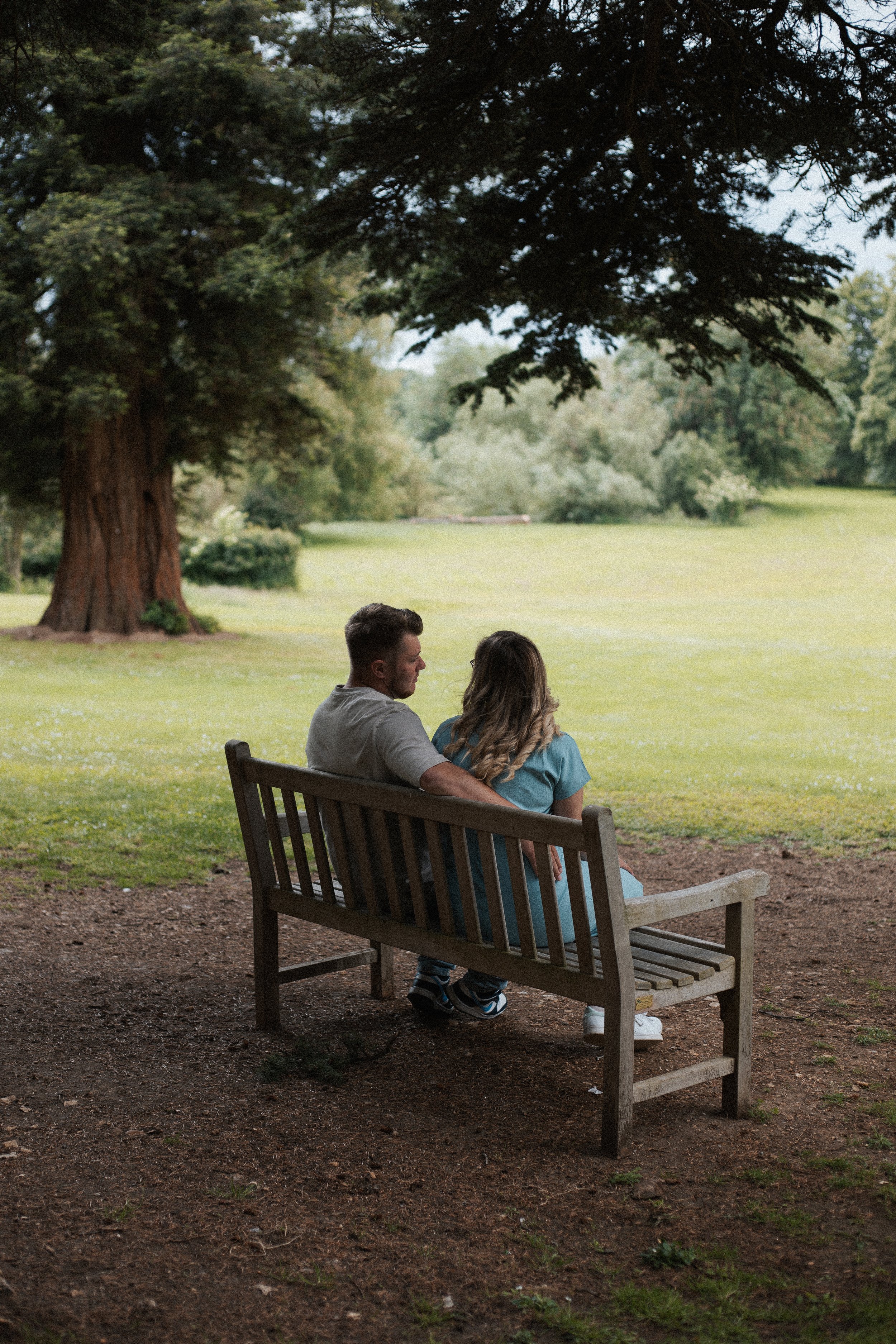 A young couple sitting on a wooden park bench under a large tree, looking at a grassy field and distant trees.
