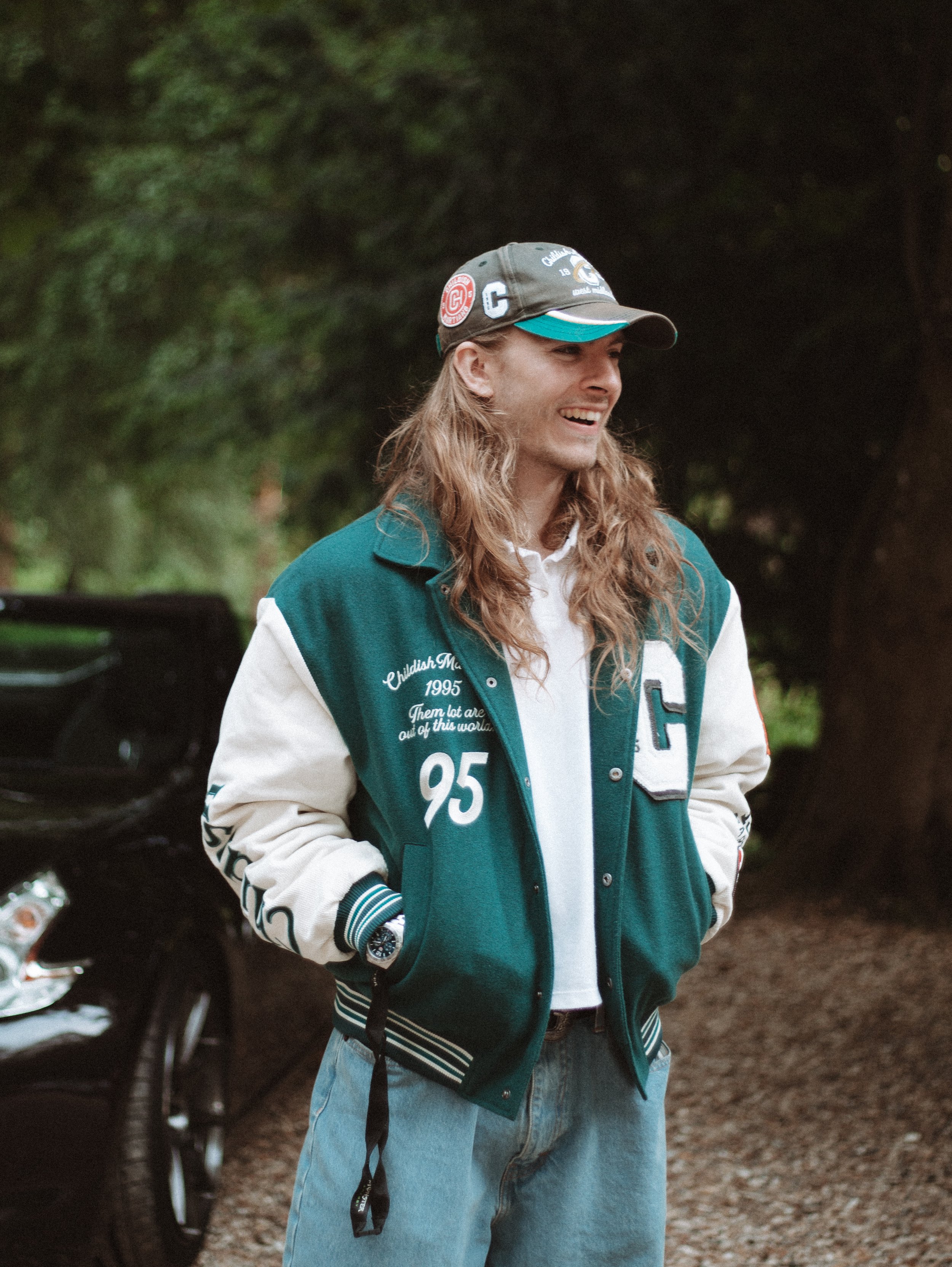 A young man with long, wavy hair wearing a teal and white varsity jacket, a baseball cap, and jeans, smiling outdoors near trees.
