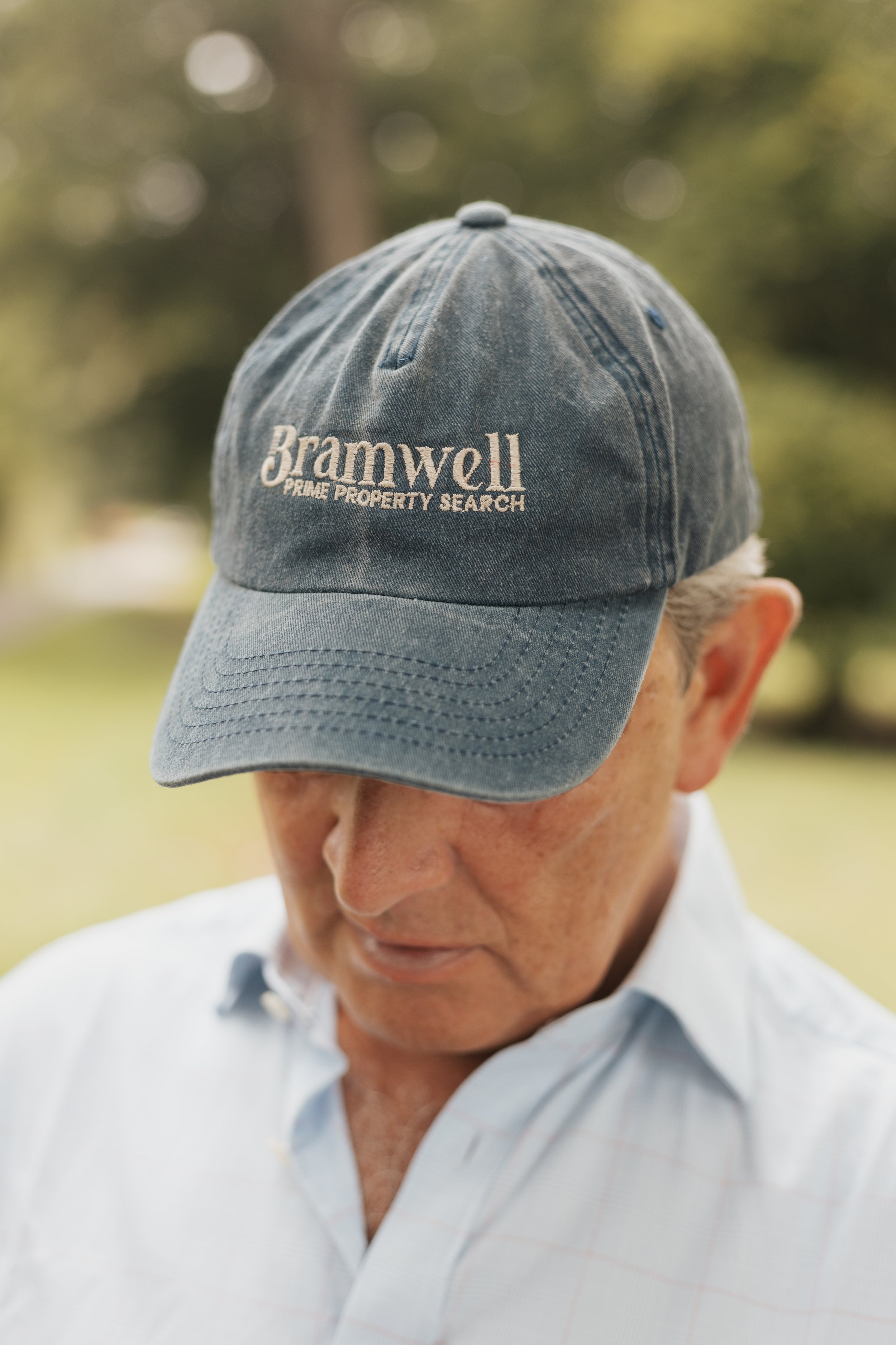 A man wearing a blue cap with the logo 'Bramwell Prime Property Search' and a light-colored collared shirt, outdoors with a blurred green background.