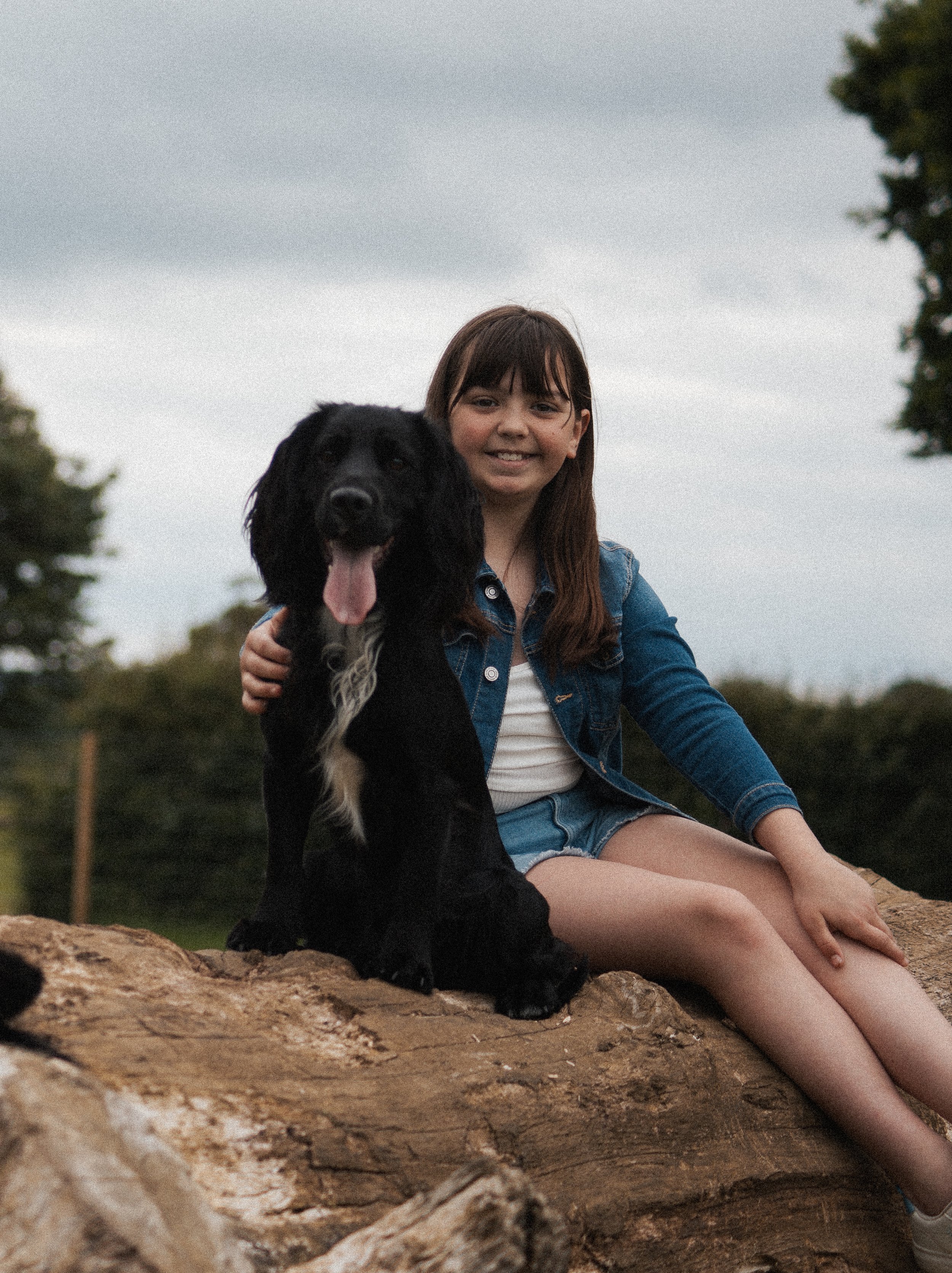 A young girl sitting on a log outdoors with a black dog, both smiling at the camera.