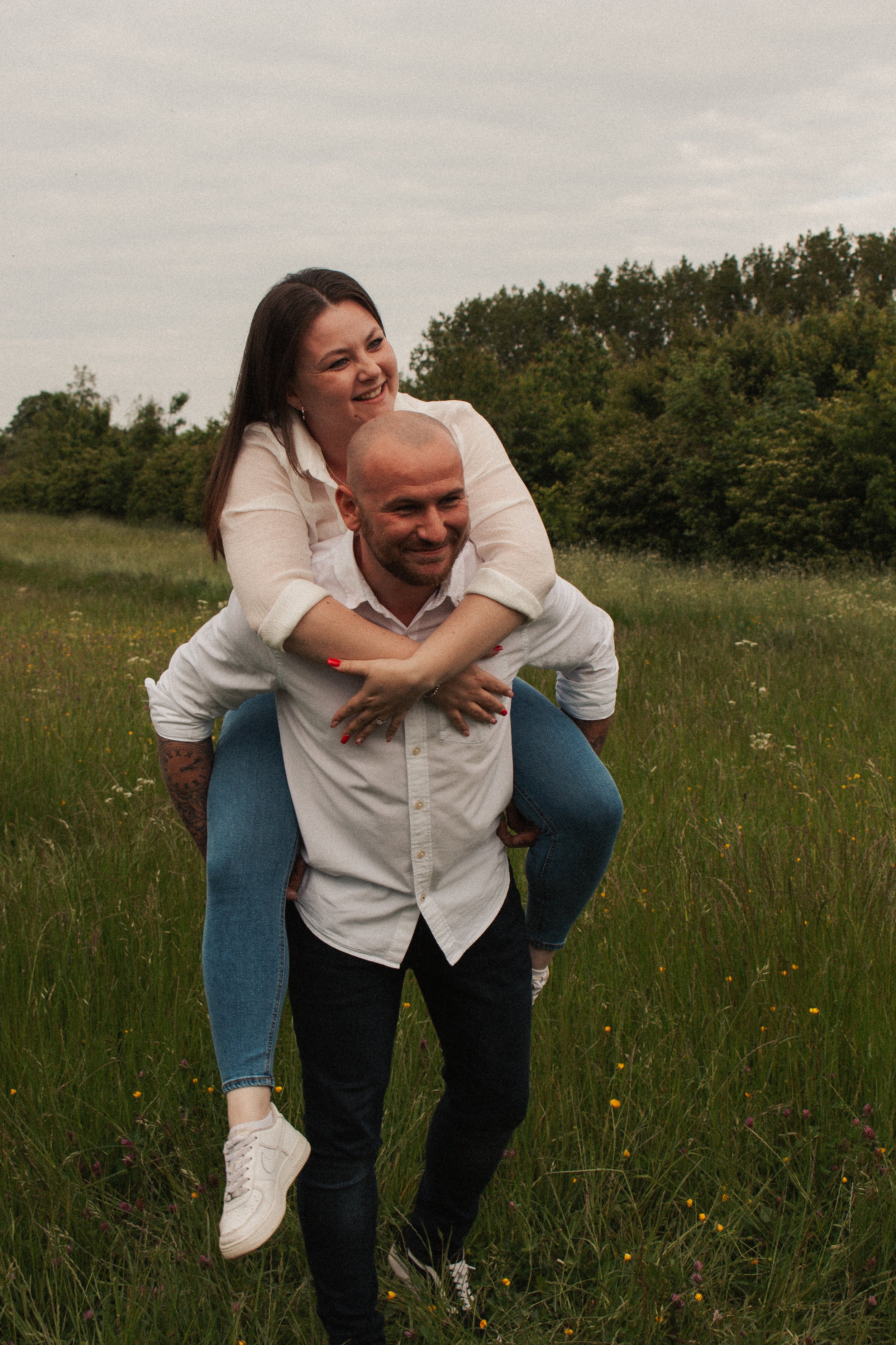 A couple is outdoors in a grassy field, with the woman on the man's back playfully smiling, and the man smiling as they enjoy a playful moment together.
