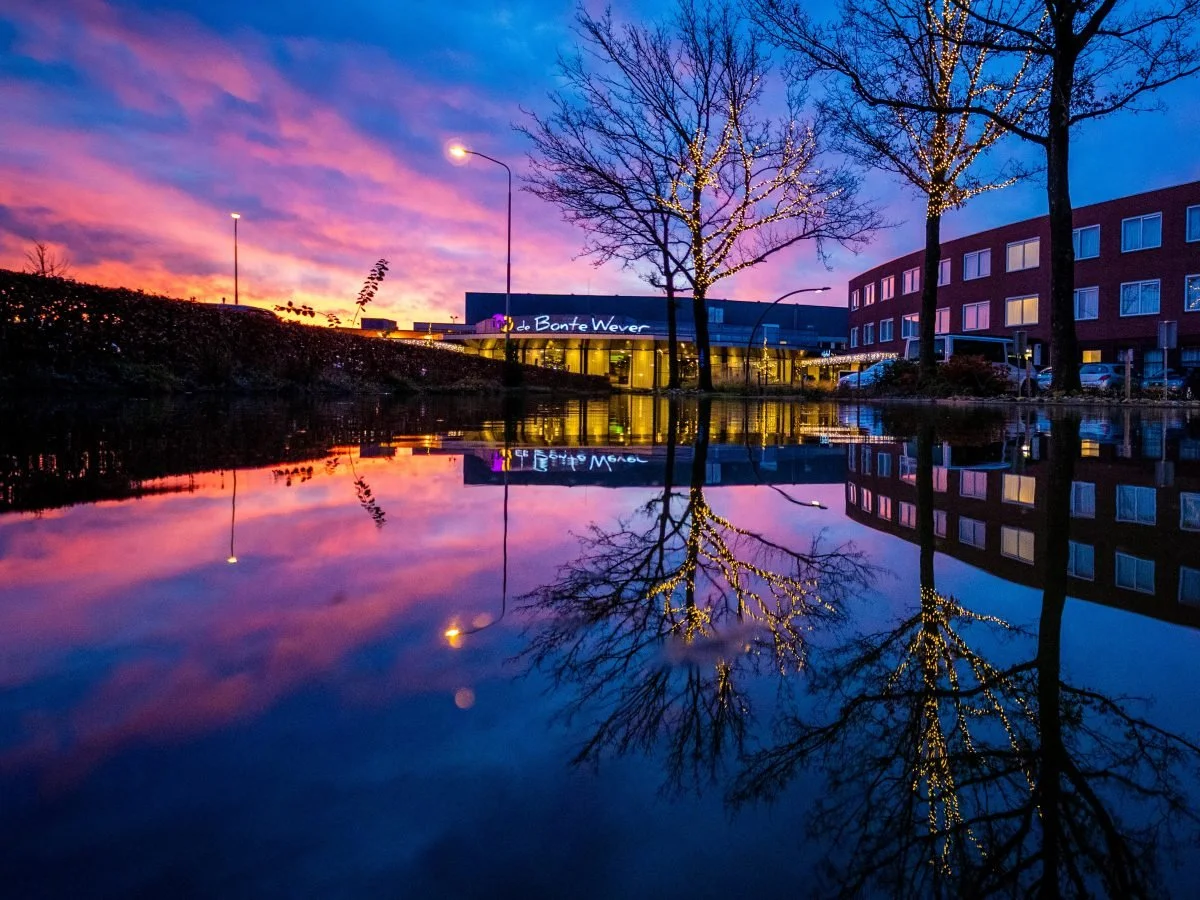 Weerspiegeling van gebouwen en bomen met sfeervolle lichtjes in het water, onder een kleurrijke avondlucht. Een rustgevend stadsbeeld vol sfeer en warmte.