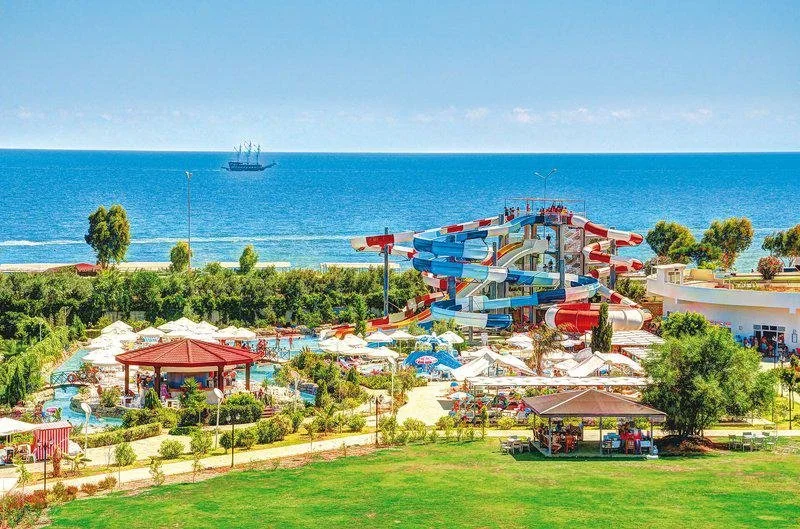 A large water park with a big colorful water slide near the ocean, surrounded by umbrellas and benches, with a pirate ship in the sea in the background.