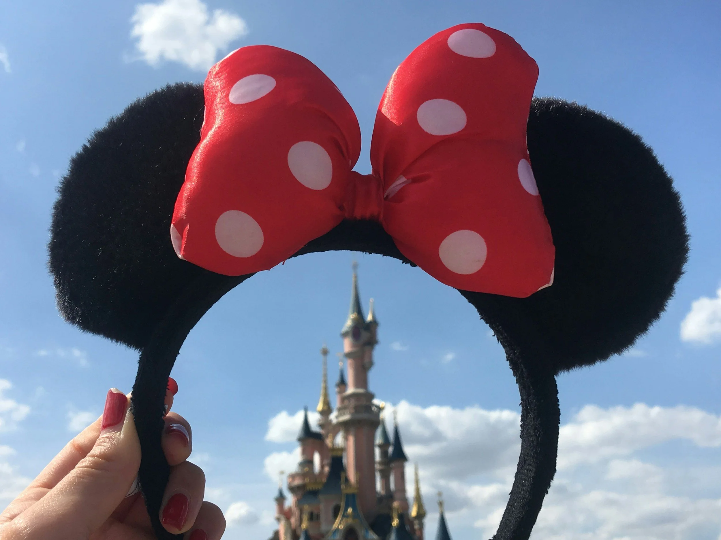 Person holding Minnie Mouse ears with a large pink bow with white polka dots at Disney theme park, Cinderella's castle in the background under a partly cloudy sky.