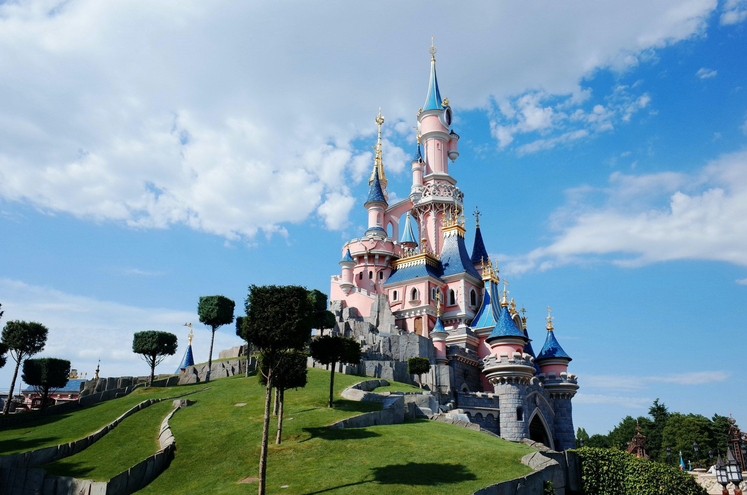 A pink and blue castle with turrets, located on a green grassy hill with trimmed trees, under a partly cloudy blue sky.