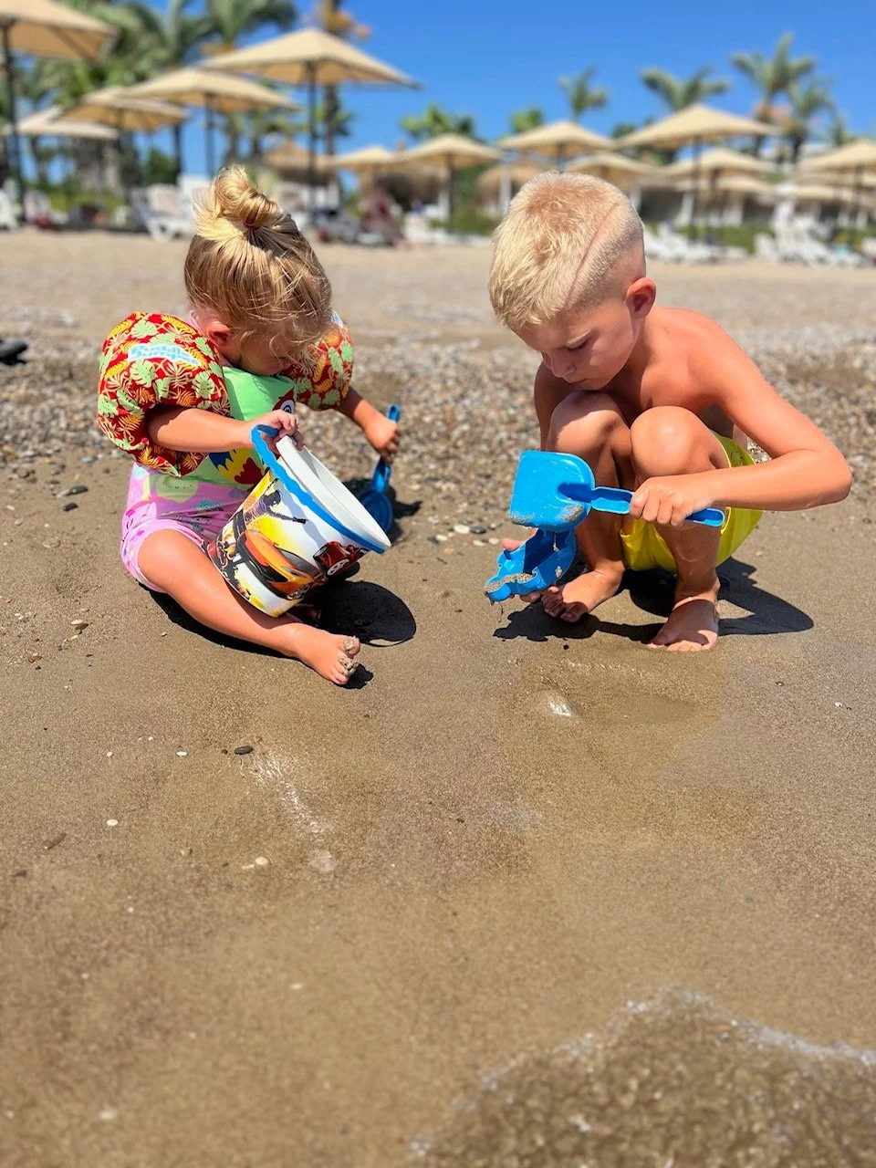 Two children, a girl and a boy, playing with toy buckets and shovels on a sandy beach under a bright blue sky with beach umbrellas in the background.