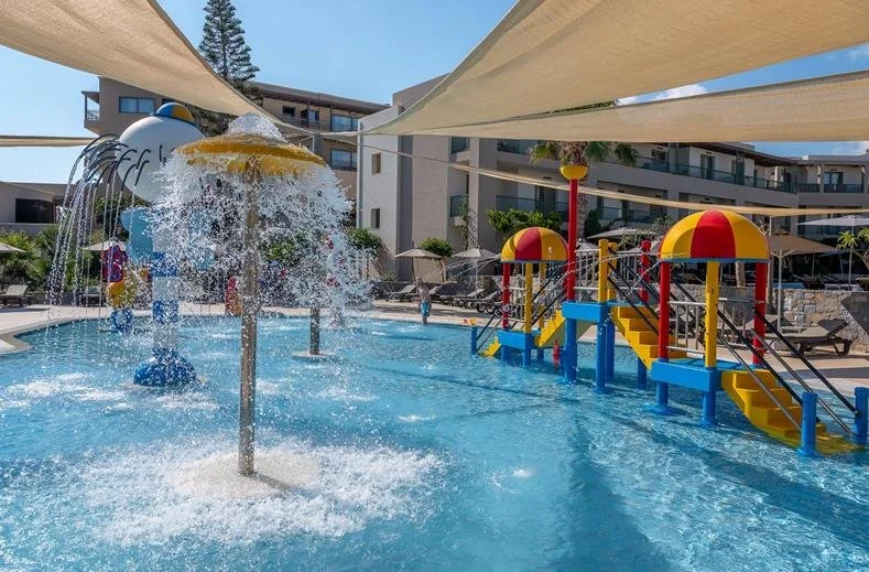 Children's water play area with splash features and small colorful structures at a resort pool, shaded by large umbrellas.