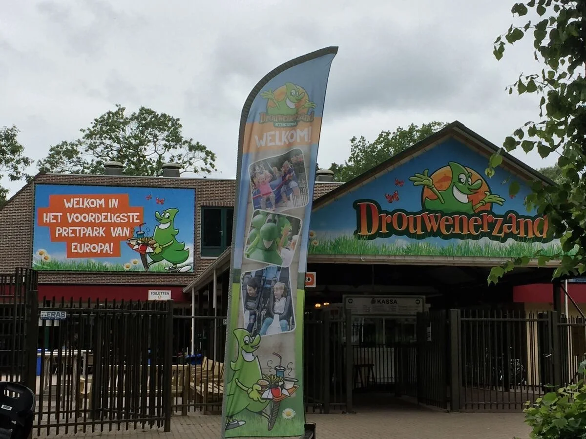 Entrance to Drouwenerzand amusement park featuring a blue sign with the park's name and a cartoon green dragon holding a tray with food, a tall vertical flag with images of park attractions and the cartoon dragon, and a large billboard welcoming visitors in Dutch.