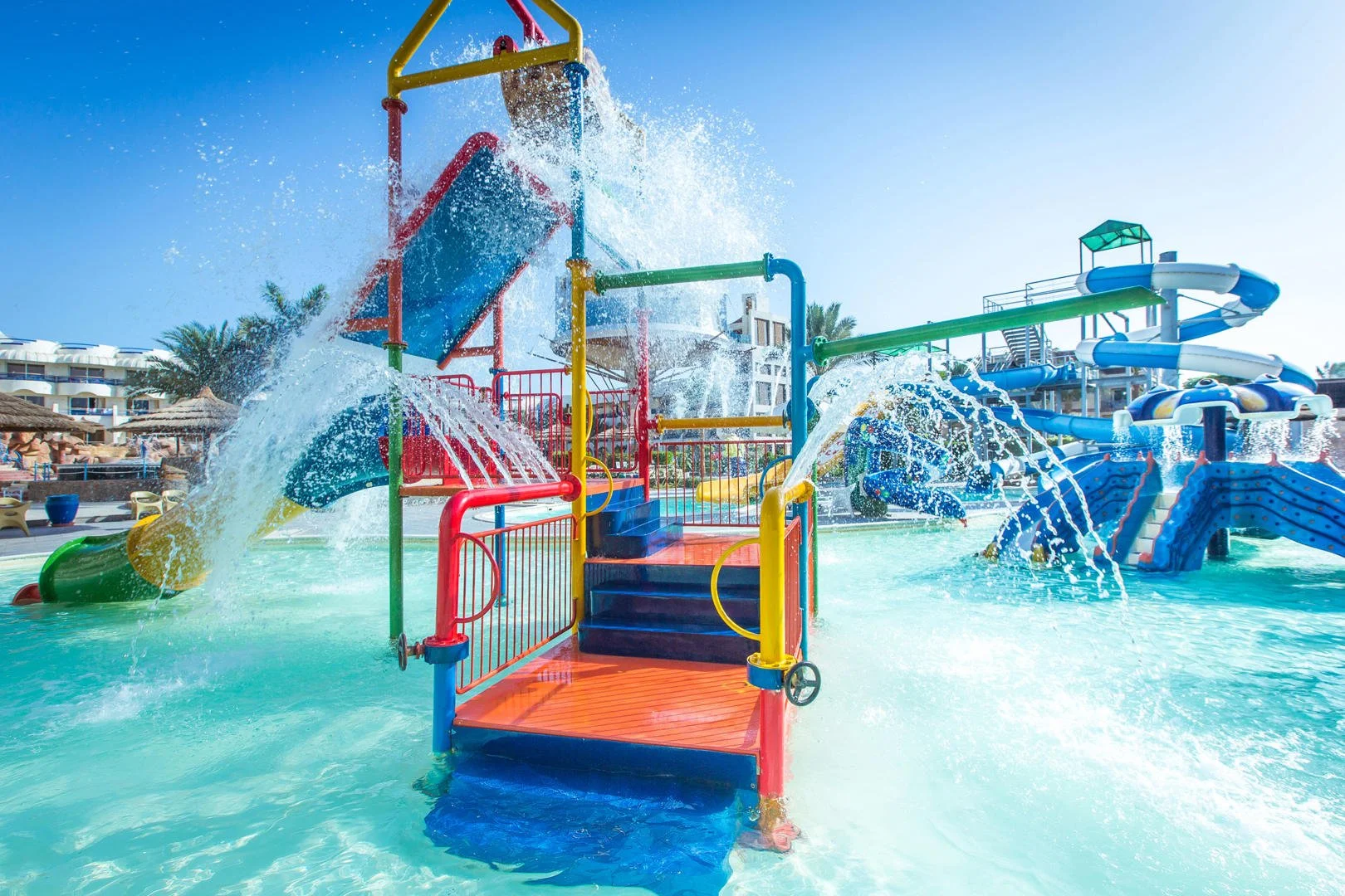 Kleurrijke waterspeeltuin met glijbanen, watersproeiers en ondiepe baden, gelegen in de buitenlucht onder een blauwe lucht. Ideaal voor jonge kinderen en perfect passend bij een kindvriendelijk vakantiepark of all inclusive resort met waterpret.