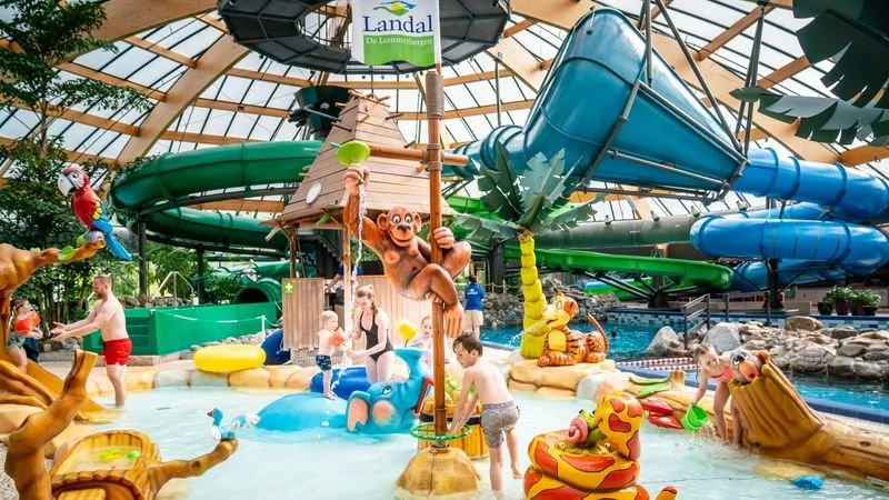 Kids playing in an indoor water park with colorful slides, water toys, and jungle-themed decorations under a glass-domed ceiling.