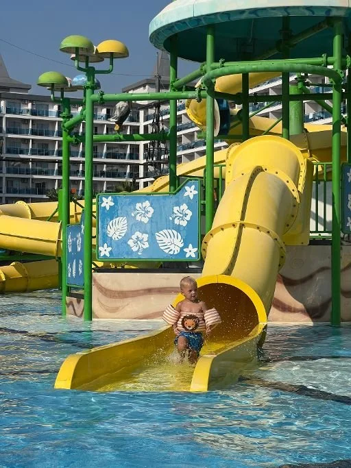 Kind dat van een gele waterglijbaan een zwembad in glijdt bij een kindvriendelijk waterpark. Op de achtergrond zijn meerdere appartementen te zien. Ideaal beeld bij een gezinsvakantie met waterpark en veel zwemplezier voor kinderen.