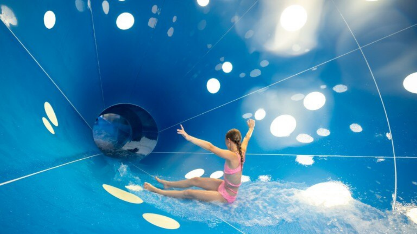Meisje in roze zwemkleding dat van een grote blauwe waterglijbaan met witte patronen glijdt. Dit beeld staat voor waterpret in een kindvriendelijk waterpark en past goed bij een gezinsvakantie met zwembaden en glijbanen.