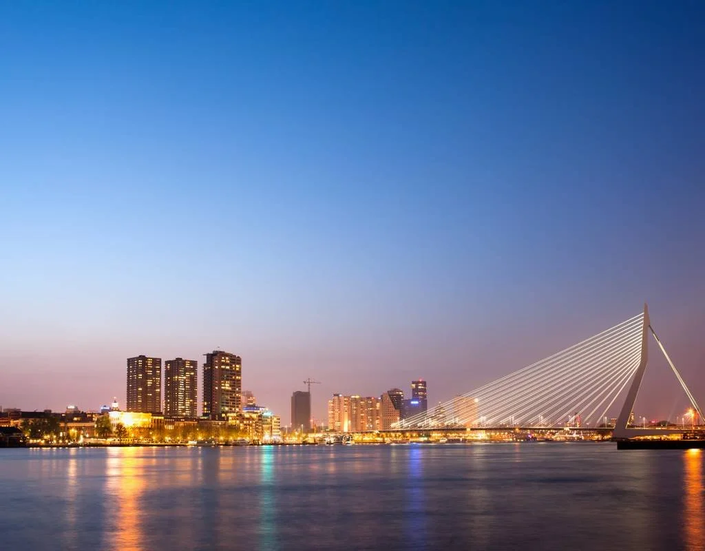 Skyline van een stad in de avond met verlichte gebouwen en een moderne brug met kabelconstructie over het water. Dit beeld past goed bij stedentrips, citytrips en vakanties in een moderne stad aan het water.