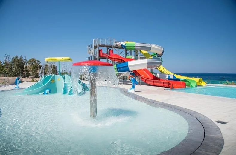 Waterpark met kleurrijke waterglijbanen, een splashgedeelte en een zwembad, gelegen vlak bij de zee op een zonnige dag. Dit beeld past perfect bij een kindvriendelijke zonvakantie, all inclusive resorts en hotels met waterparken aan zee.