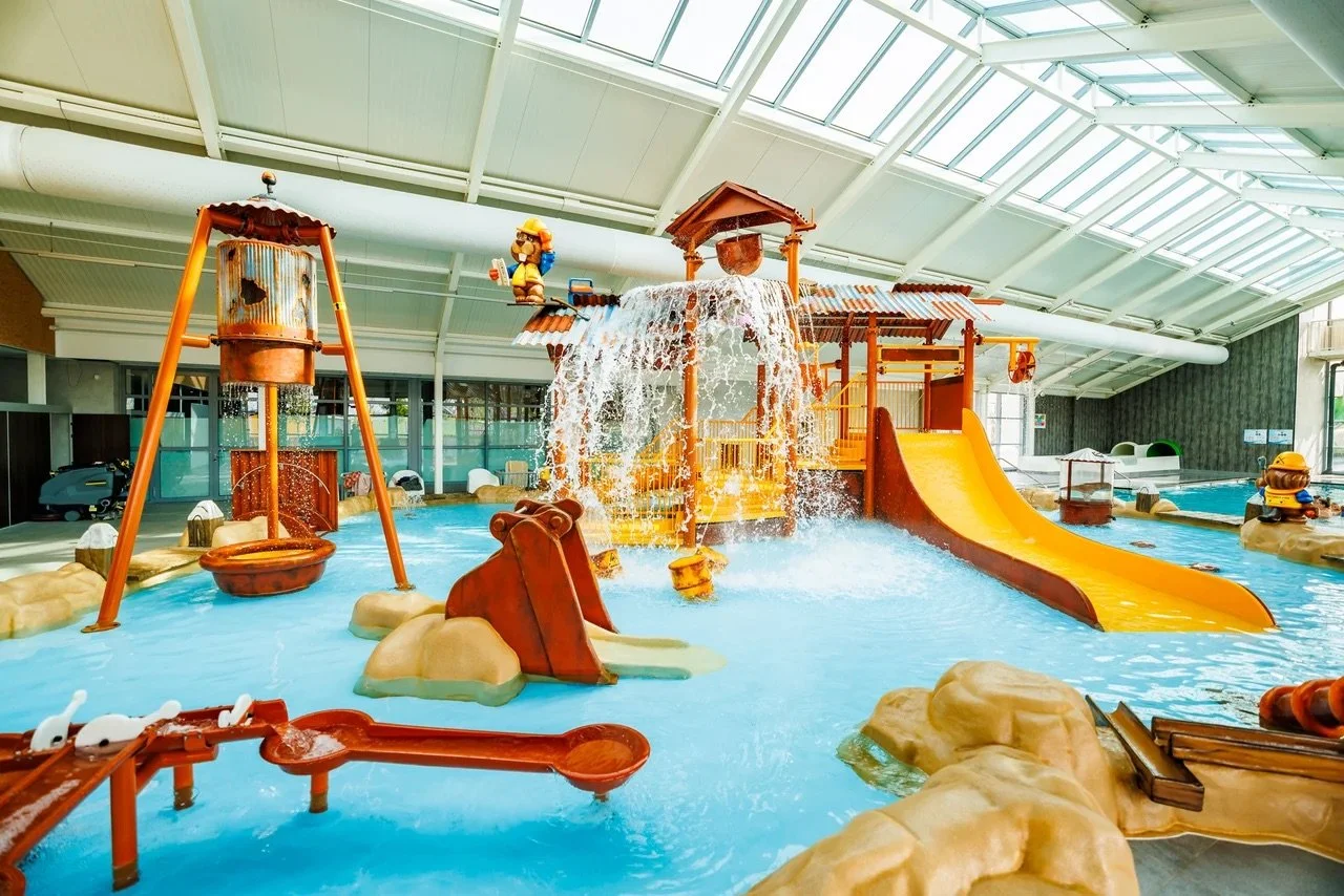 Overdekt waterpark met een grote gele glijbaan, watertoestellen en kinderen die spelen in een ondiep zwembad onder een glazen dak.