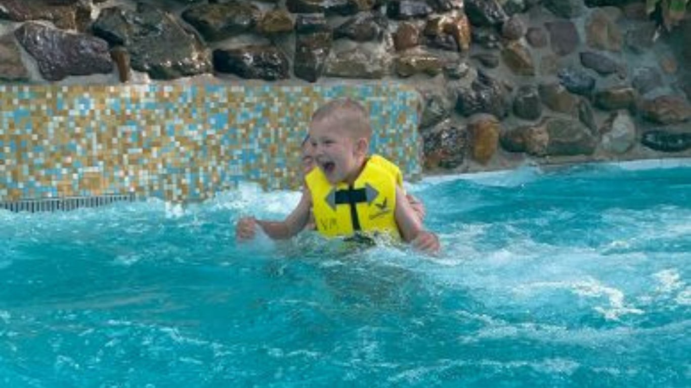 Jonge jongen met een geel zwemvest die lachend speelt in een zwembad met helder blauw water.Op de achtergrond zijn een betegelde wand en rotsen te zien. Dit beeld past perfect bij een kindvriendelijke vakantie met zwembaden en waterpret voor gezinnen