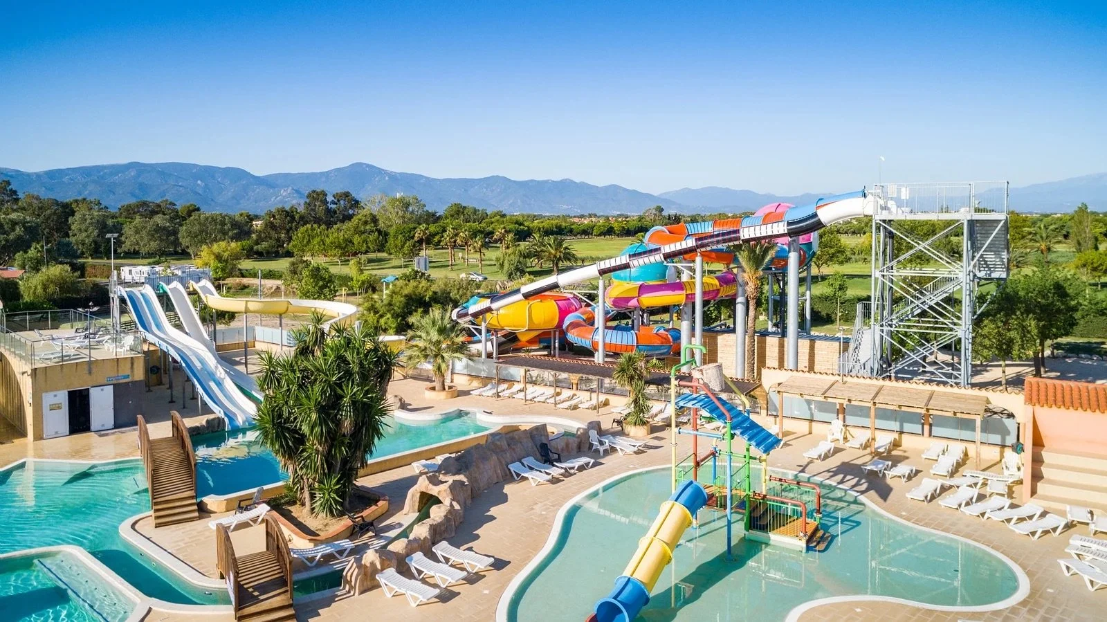 A water park with multiple water slides, a swimming pool, lounge chairs, palm trees, and a mountain range in the background on a sunny day.